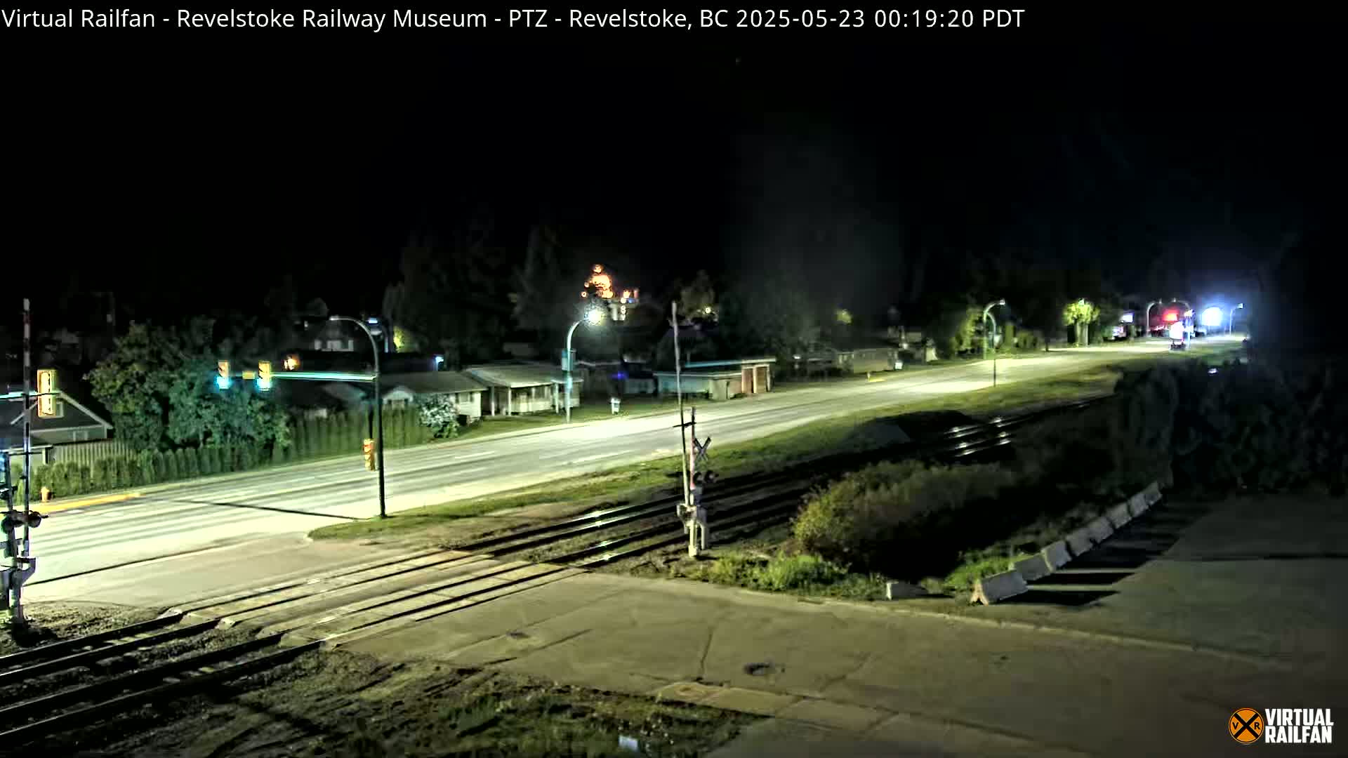 A nighttime view of a road and railway crossing, with streetlights illuminating a mostly empty roadway and houses in the background.