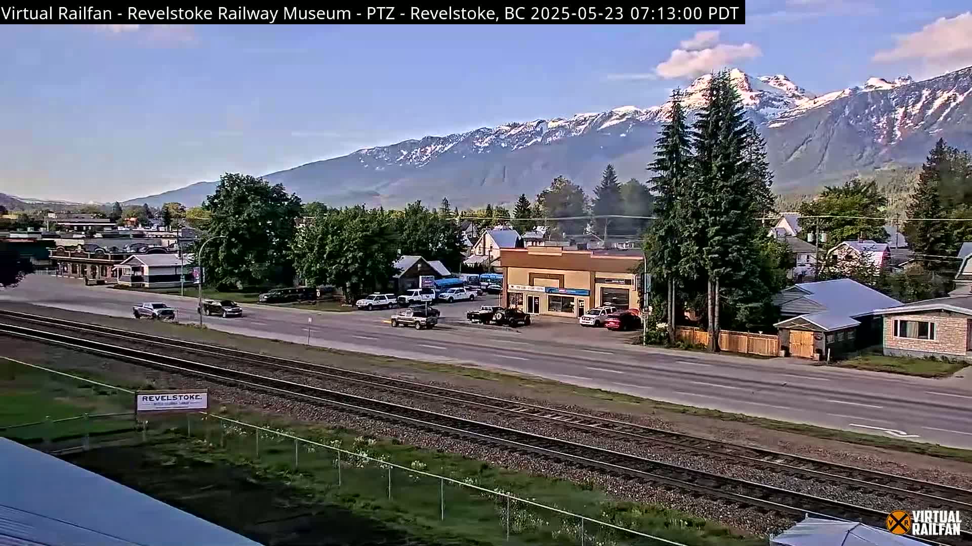 A clear, sunny day reveals a small town nestled in a valley, with snow-capped mountains in the background and a railway track running through the foreground.
