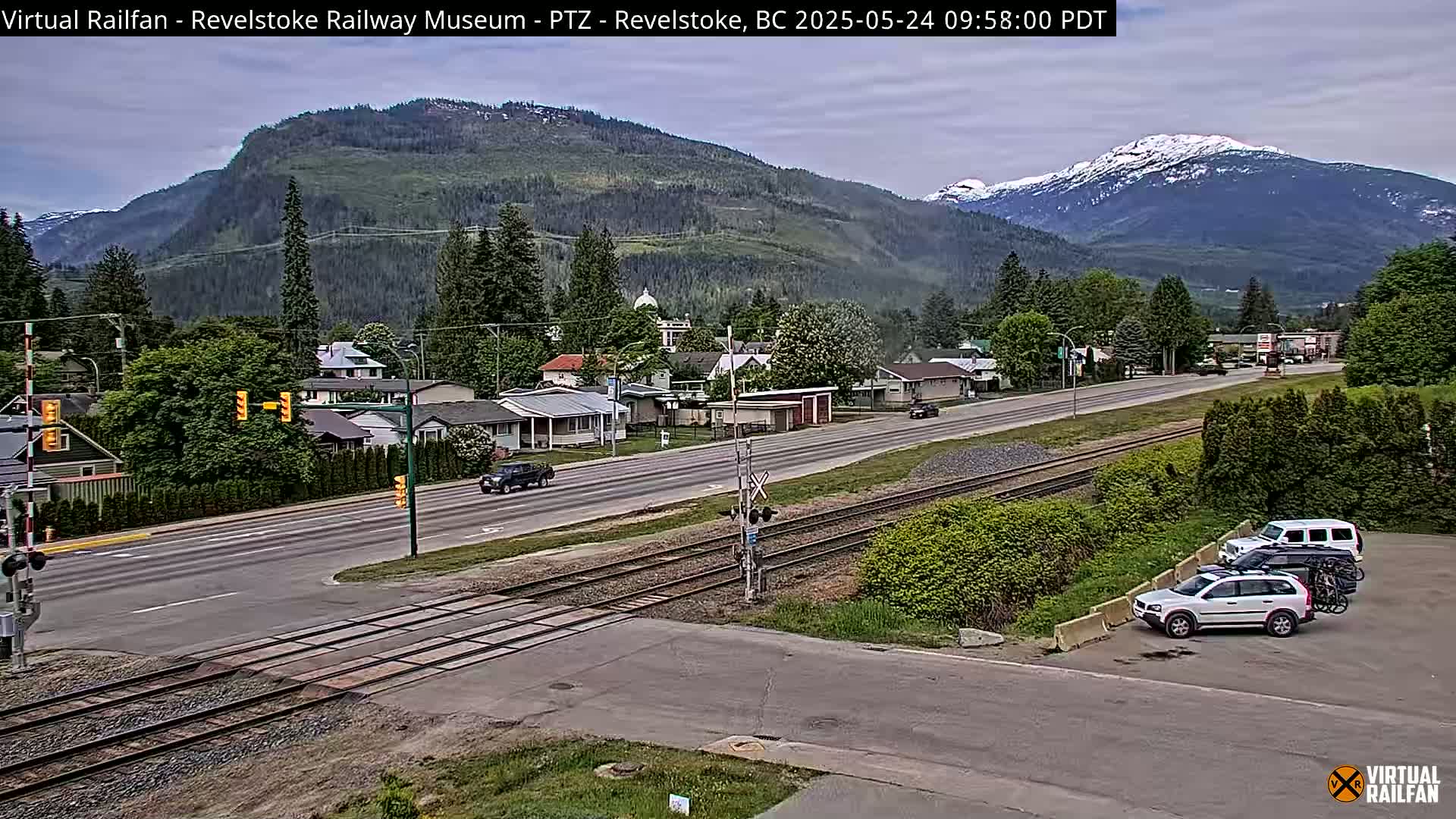 A partly cloudy day shows a town nestled in a valley between mountains, some of which have snow-capped peaks, with a railway line running through the town and cars parked and driving on roads.