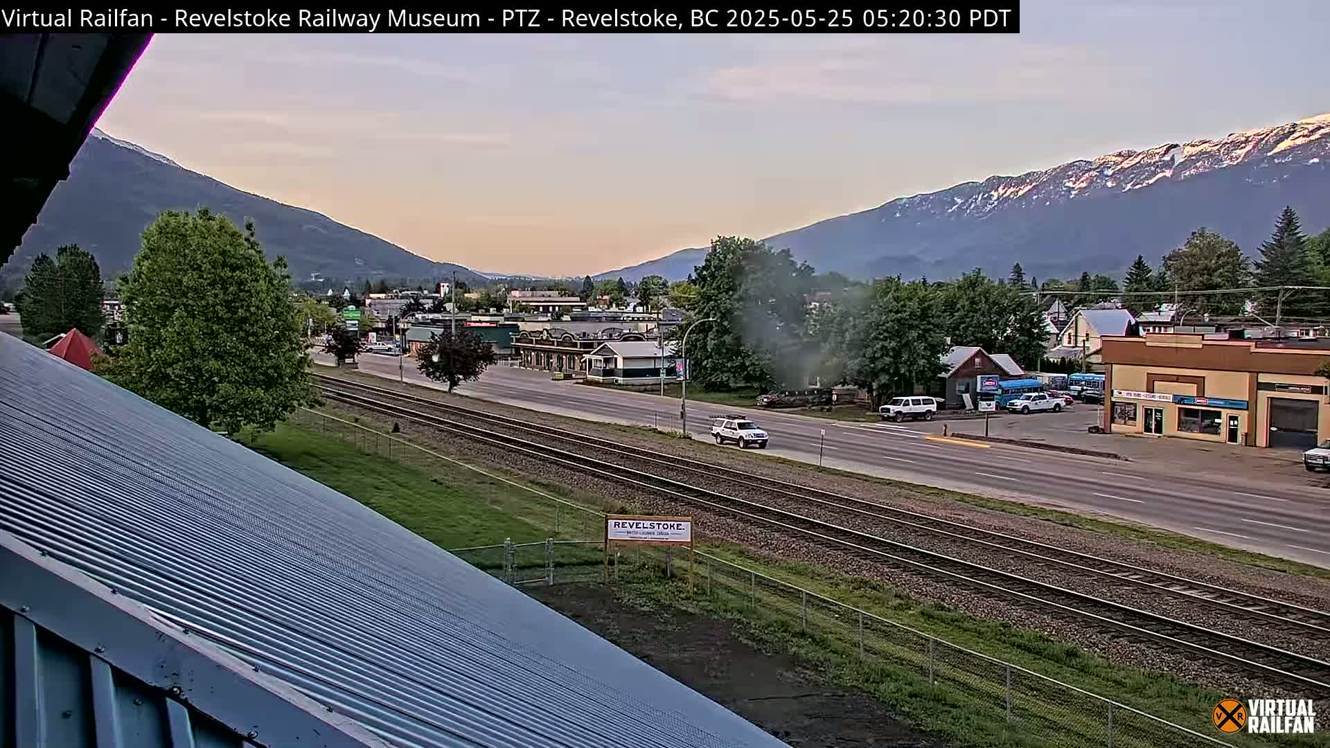 A view from a rooftop shows a town nestled in a valley between snow-capped mountains under a twilight sky, with railroad tracks running parallel to a road.