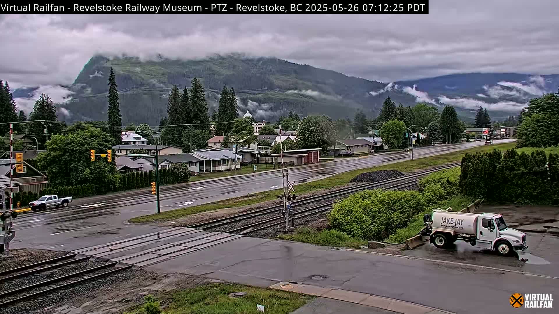 A wet town with a railway crossing, flanked by trees and a mountain shrouded in clouds.