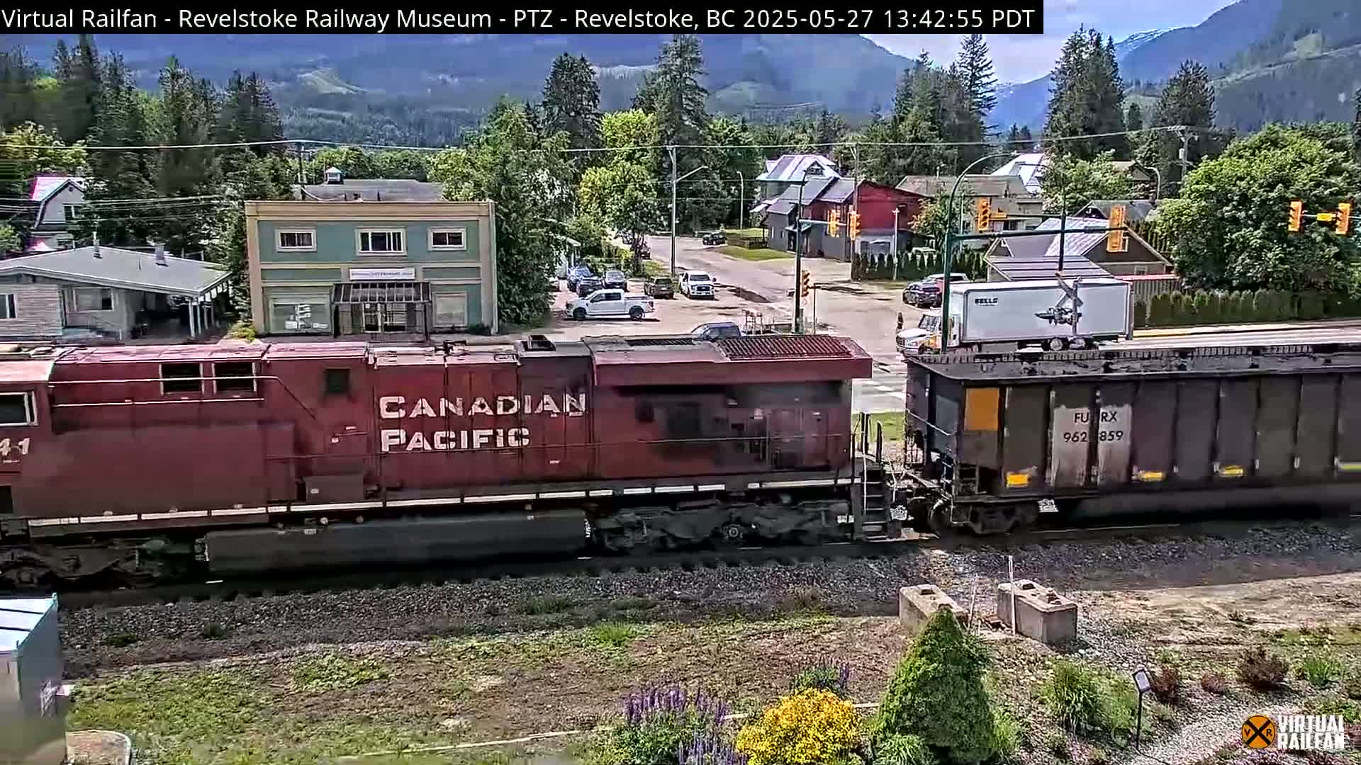A Canadian Pacific Railway train passes through a small town on a sunny day, with mountains in the background.