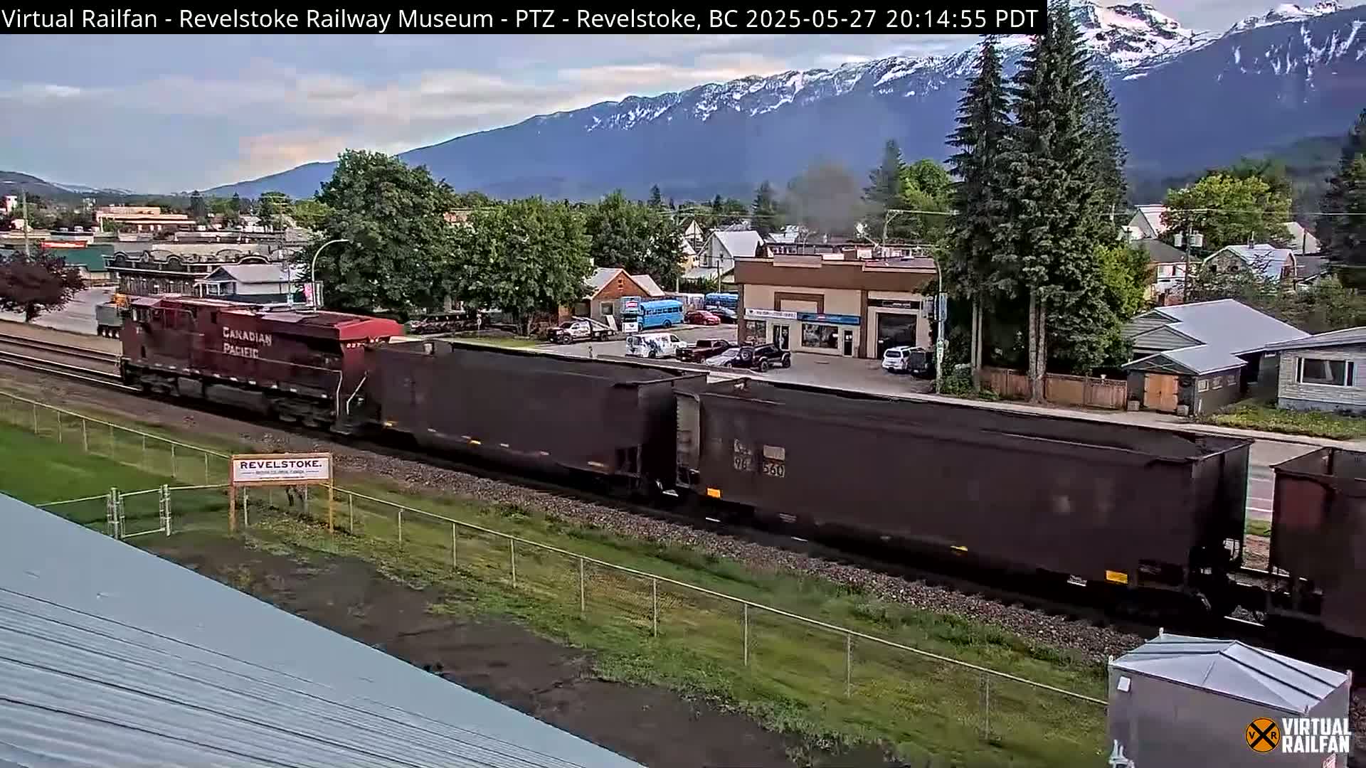 A long freight train, pulled by a red locomotive, passes through a town nestled in a valley beneath snow-capped mountains on a partly cloudy evening.