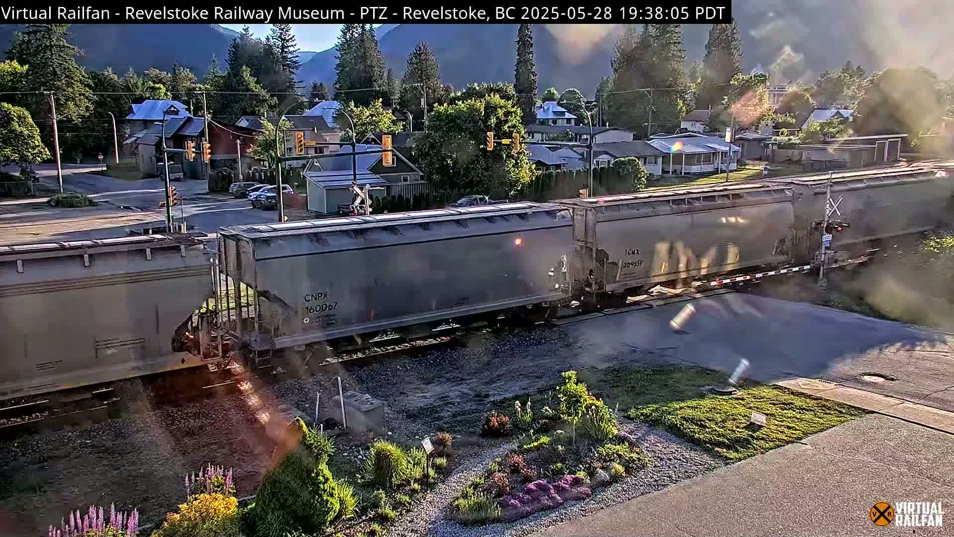 A long freight train passes a residential street crossing under partly cloudy evening skies.