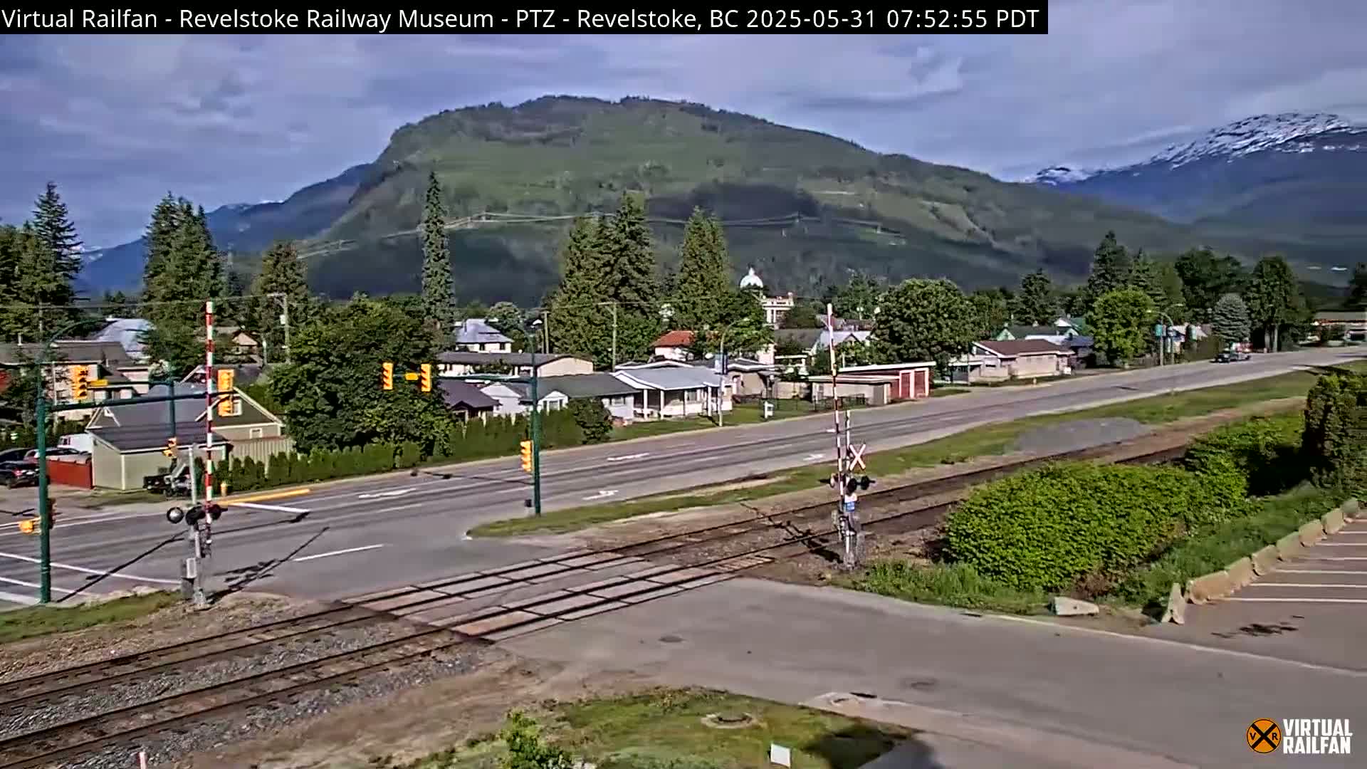 A partly sunny day reveals a town nestled in a valley beneath a mountain range, with a road crossing railway tracks in the foreground.