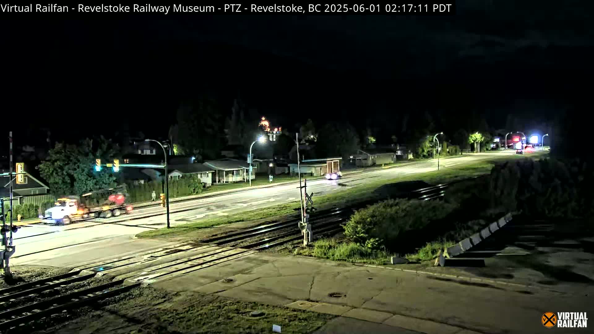A nighttime view of a road intersecting railroad tracks, with a few vehicles on the road and houses and trees visible in the background under a dark sky.