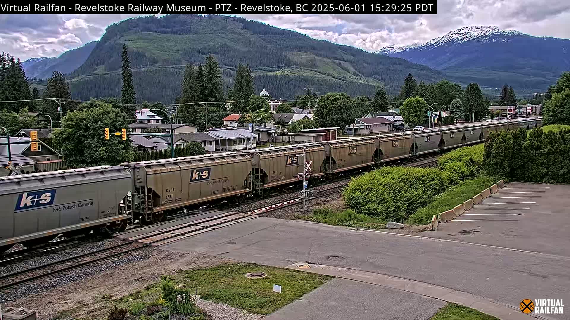 A long freight train passes through a residential area on a partly cloudy day, with mountains in the background.