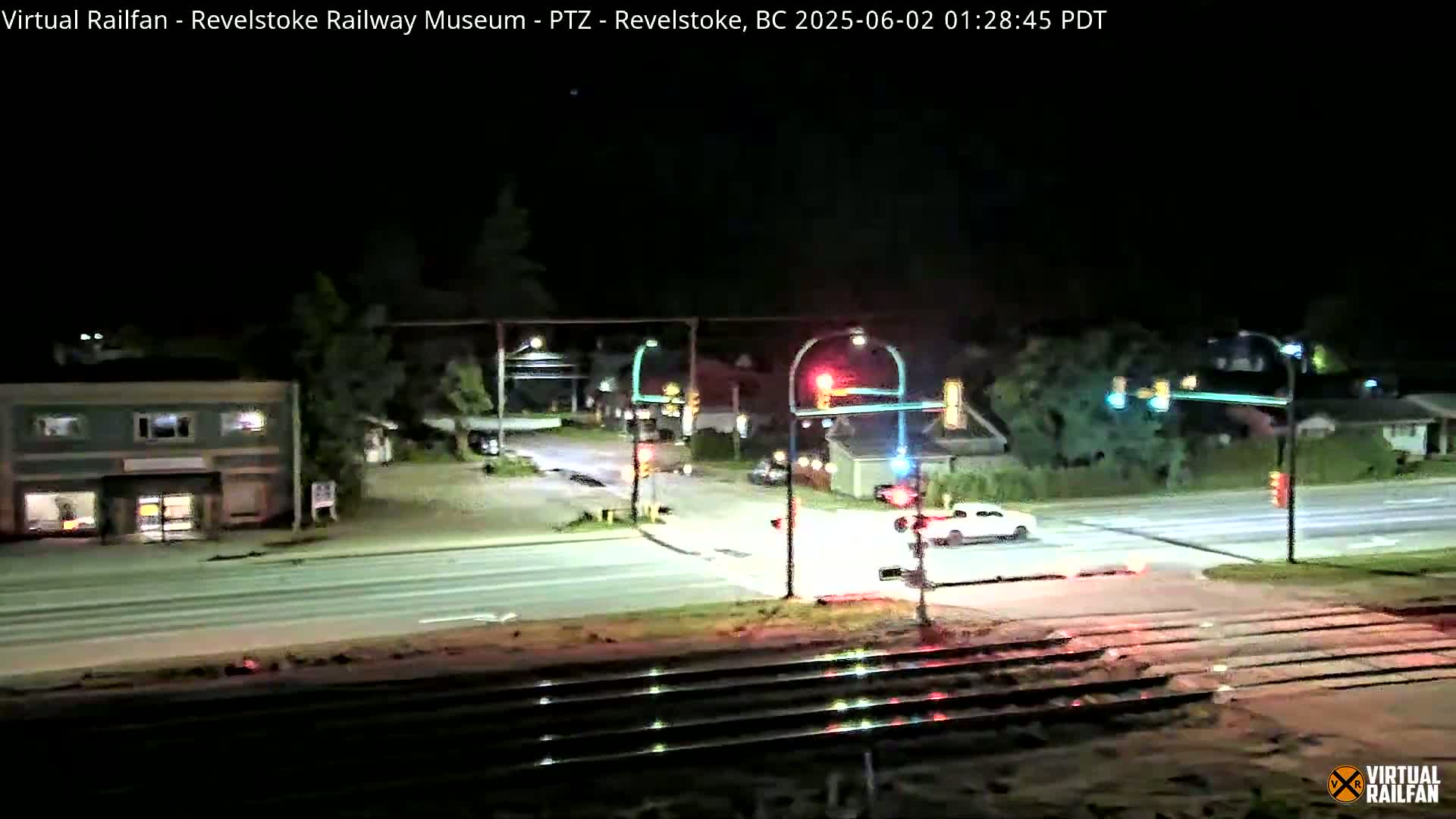 A nighttime view of a street intersection with a railway crossing, showing cars at traffic signals and a single-story building across the street.