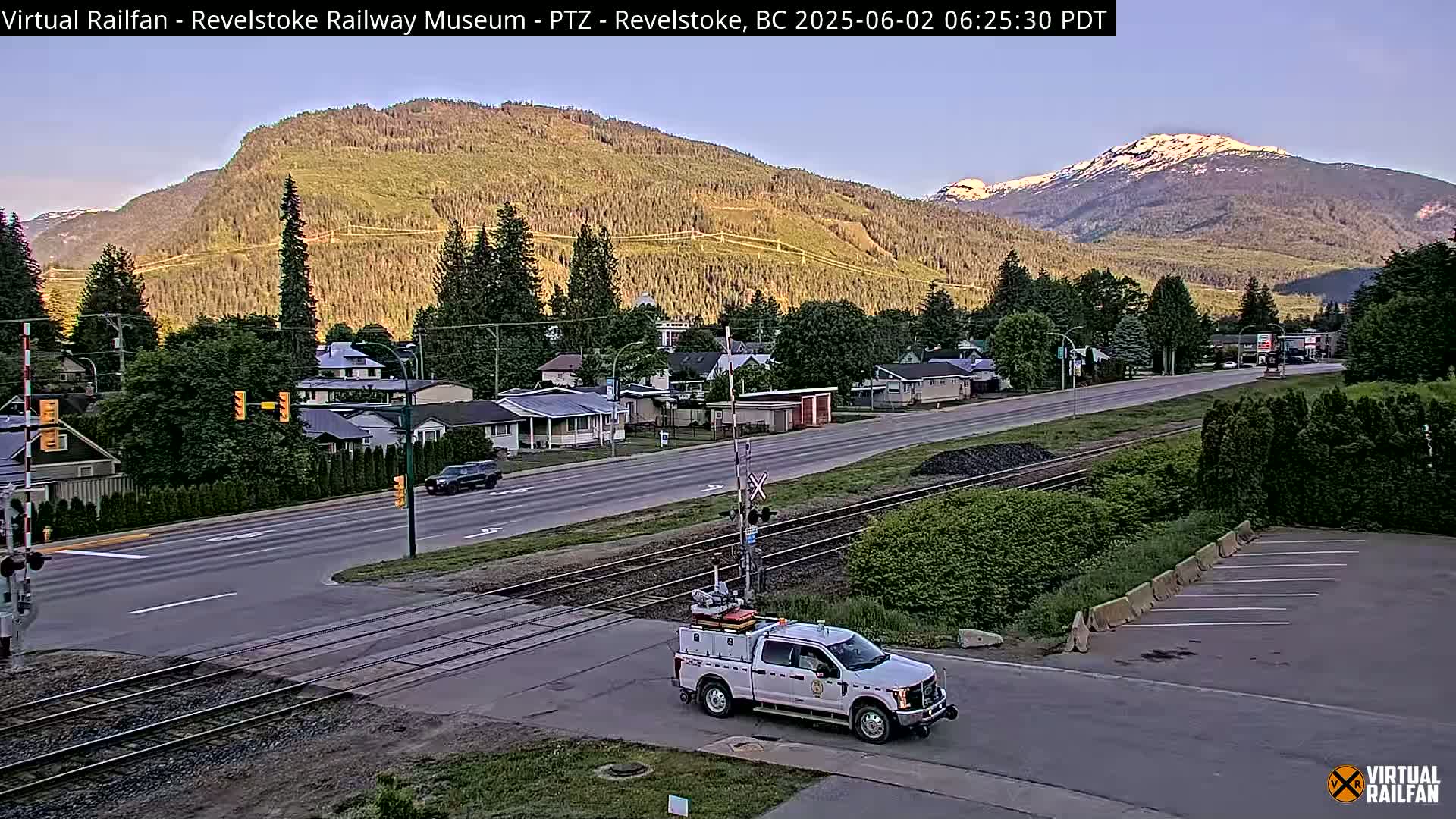 A partly sunny morning view of a small town nestled in a valley between mountains, showing a road crossing railroad tracks with a white pickup truck at the crossing.