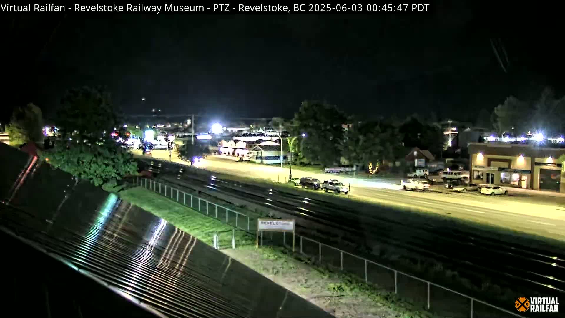 A nighttime view of a road and railway tracks, with several vehicles parked and driving, near some buildings under a clear, dark sky.