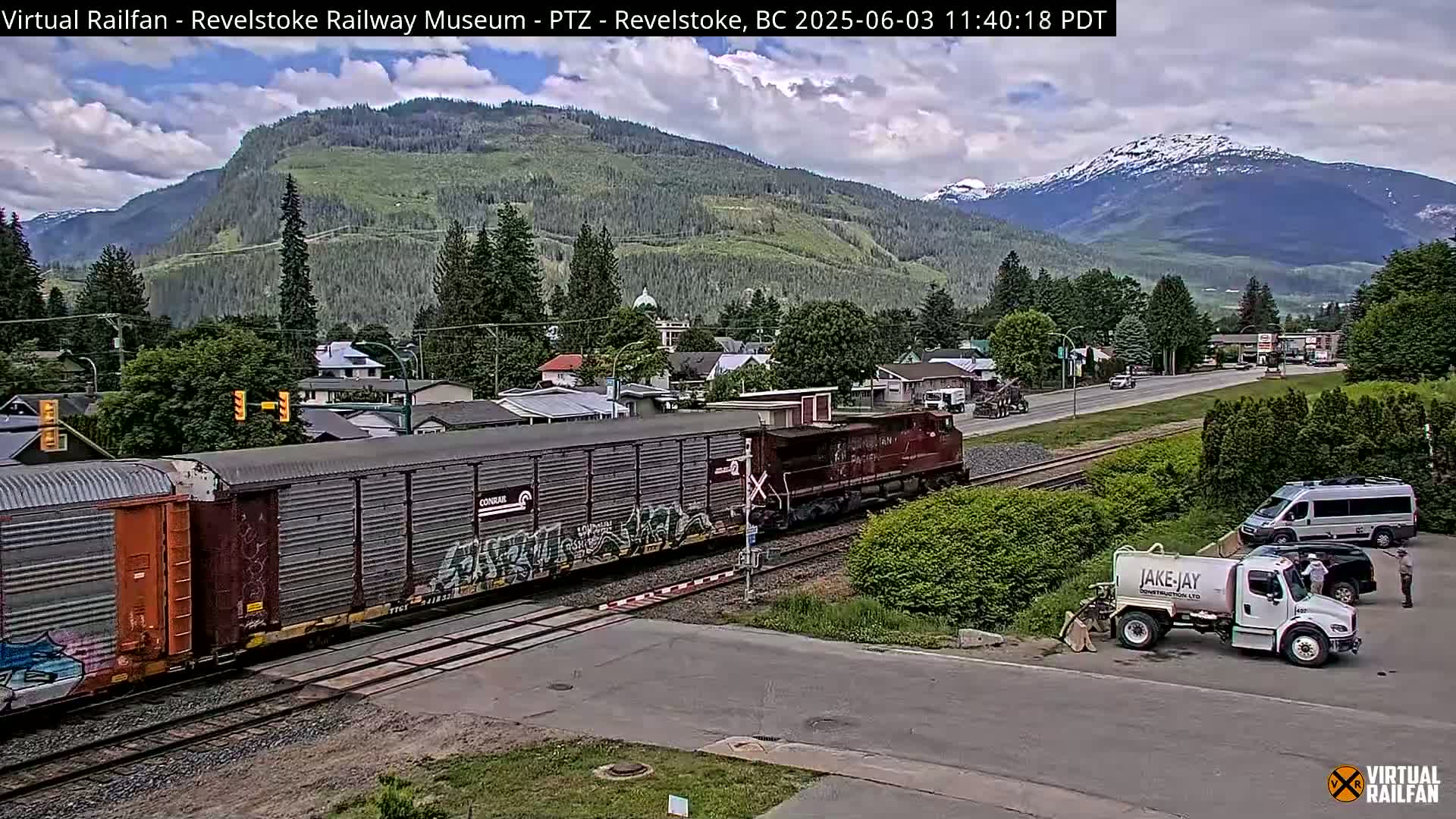 A freight train passes through a town nestled in a valley, with mountains in the background under partly cloudy skies.