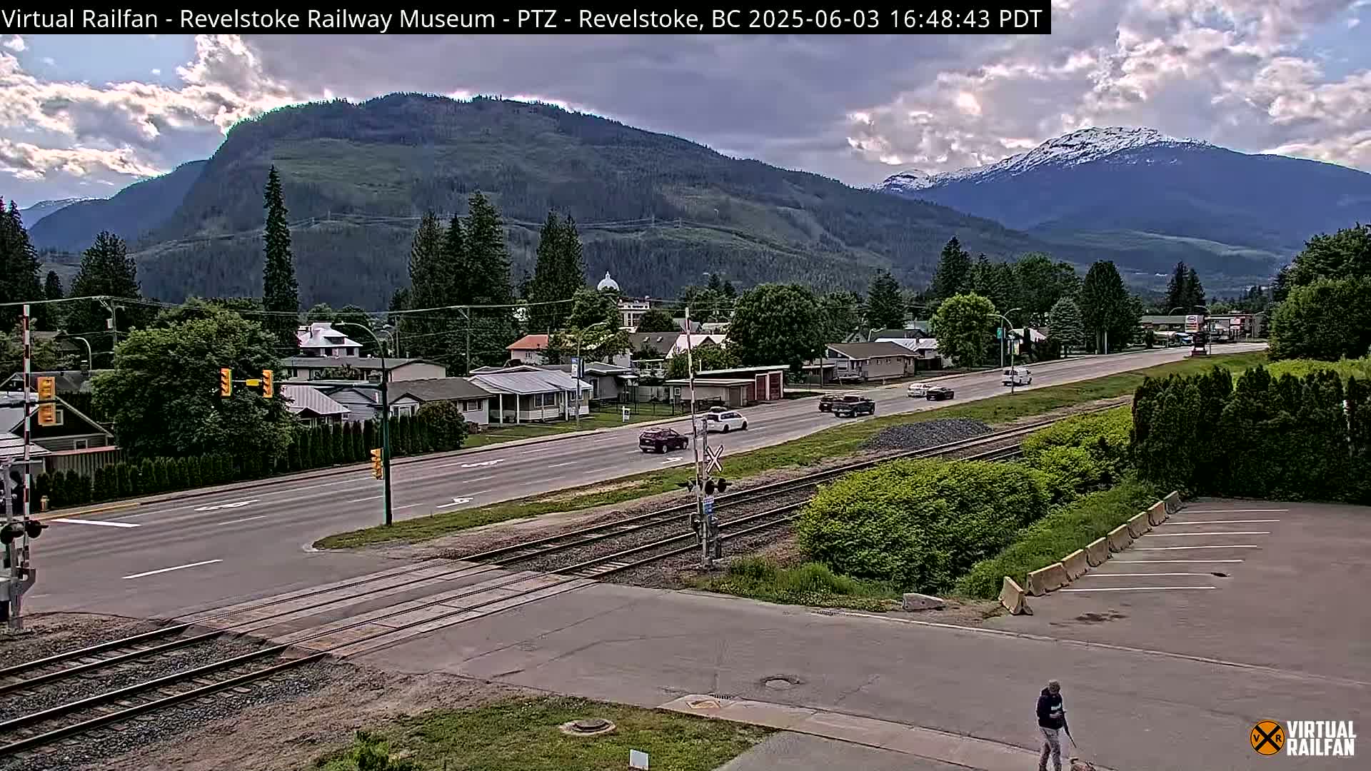 A partly cloudy day reveals a town nestled in a valley between mountains, with a road and railway tracks intersecting in the foreground.