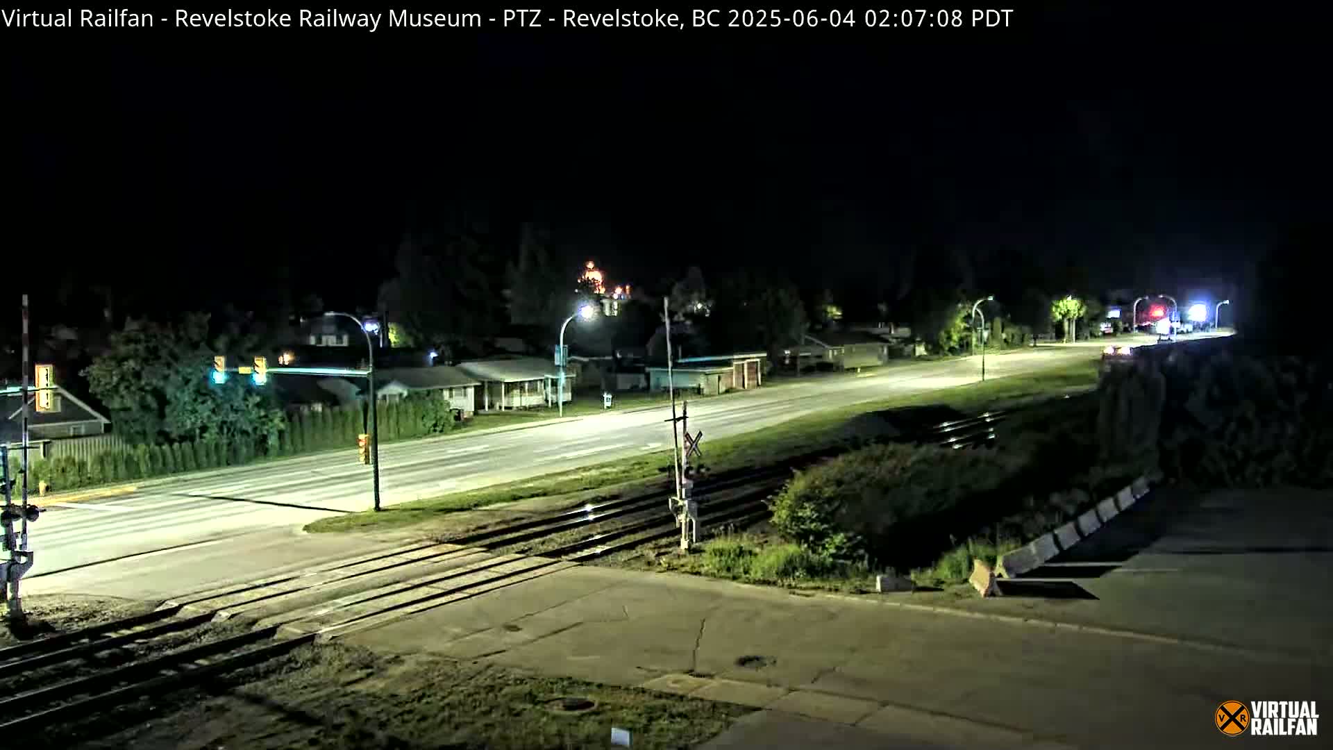 A nighttime view of a road and railway crossing, with houses and streetlights visible in the background under a clear sky.