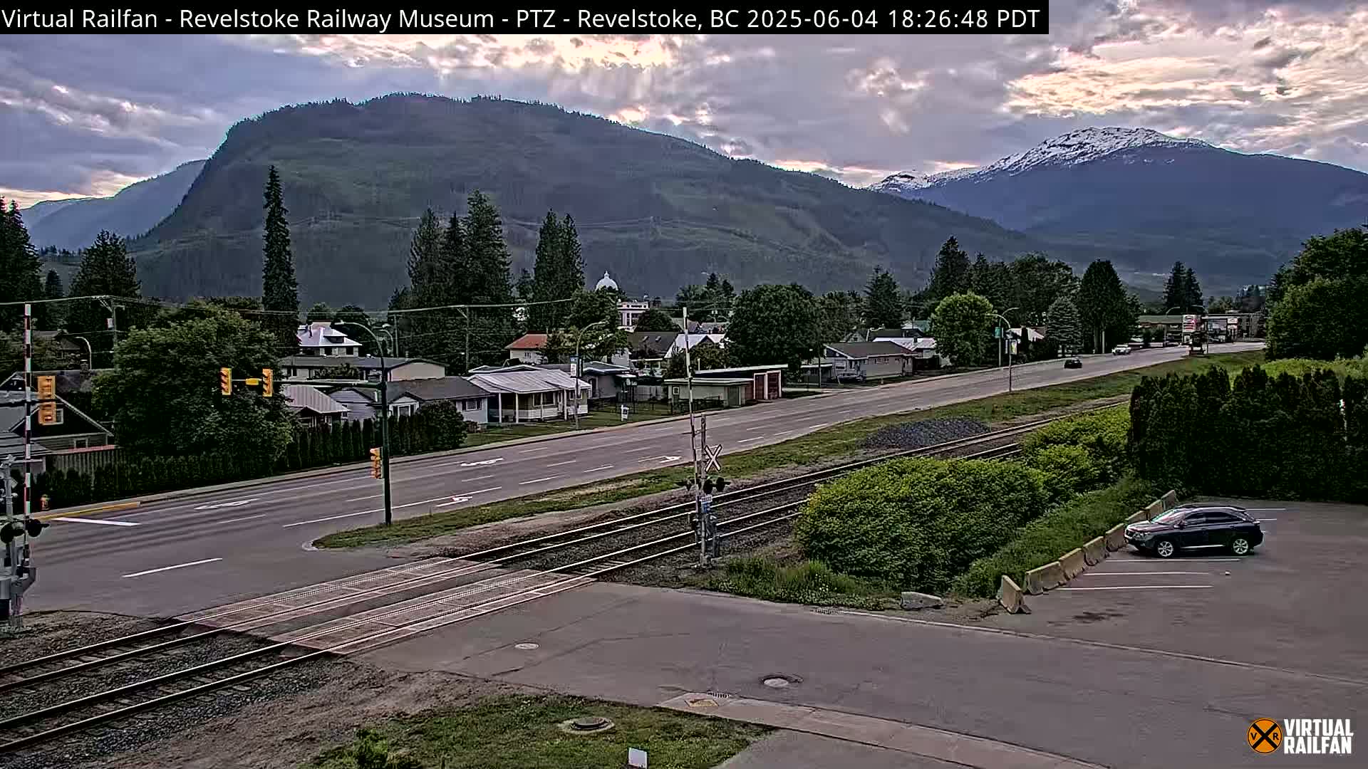 A partly cloudy late afternoon view of a town nestled in a valley between mountains, with train tracks crossing a road in the foreground.
