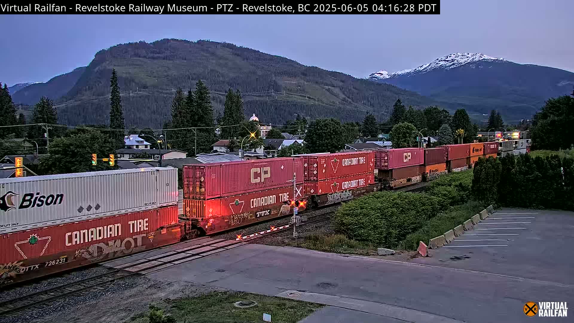 A freight train with red and white containers passes through a town nestled in a valley, with mountains in the background under a twilight sky.