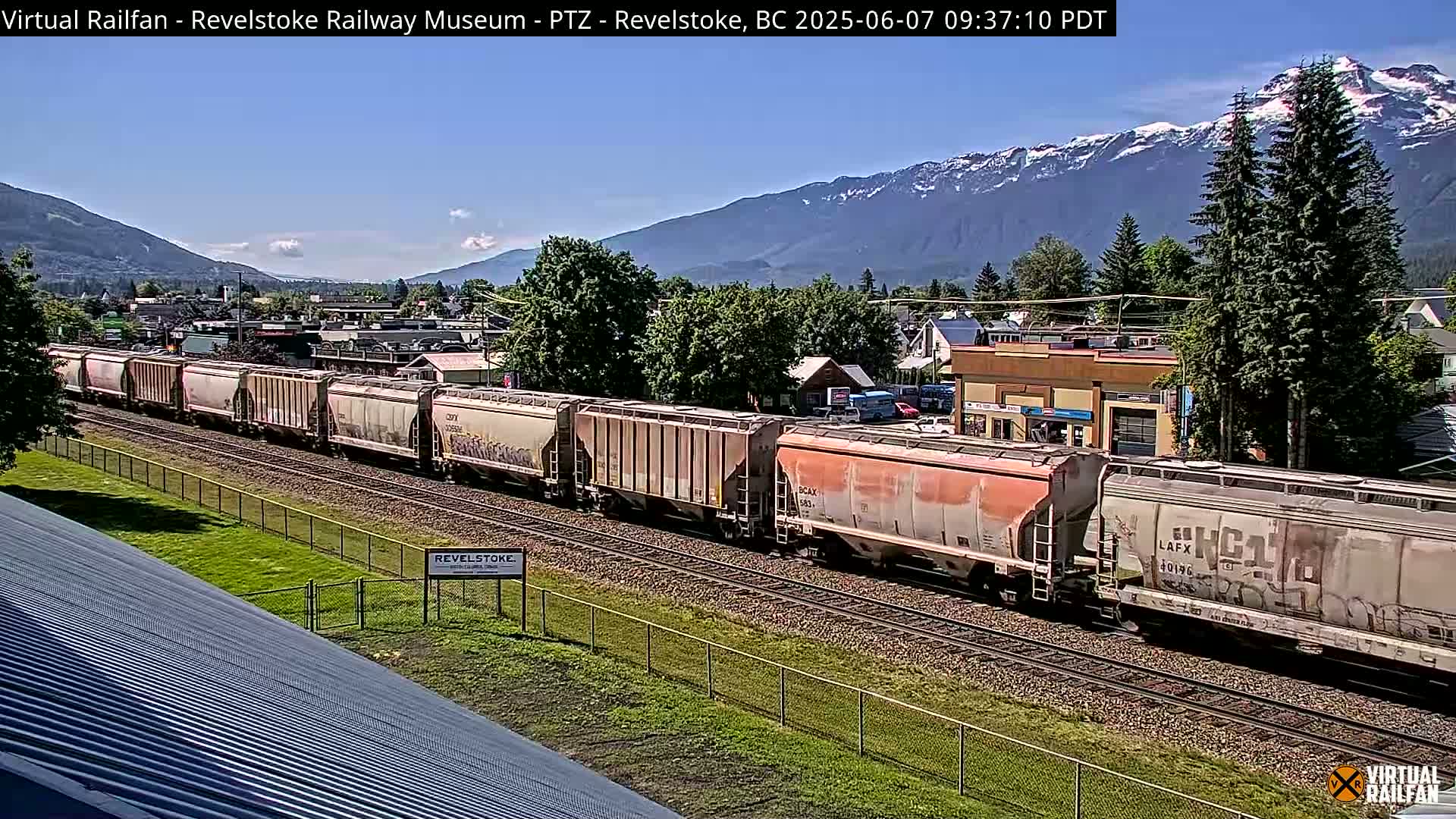 A long freight train passes through a town nestled in a valley, with snow-capped mountains visible in the background under a clear blue sky.