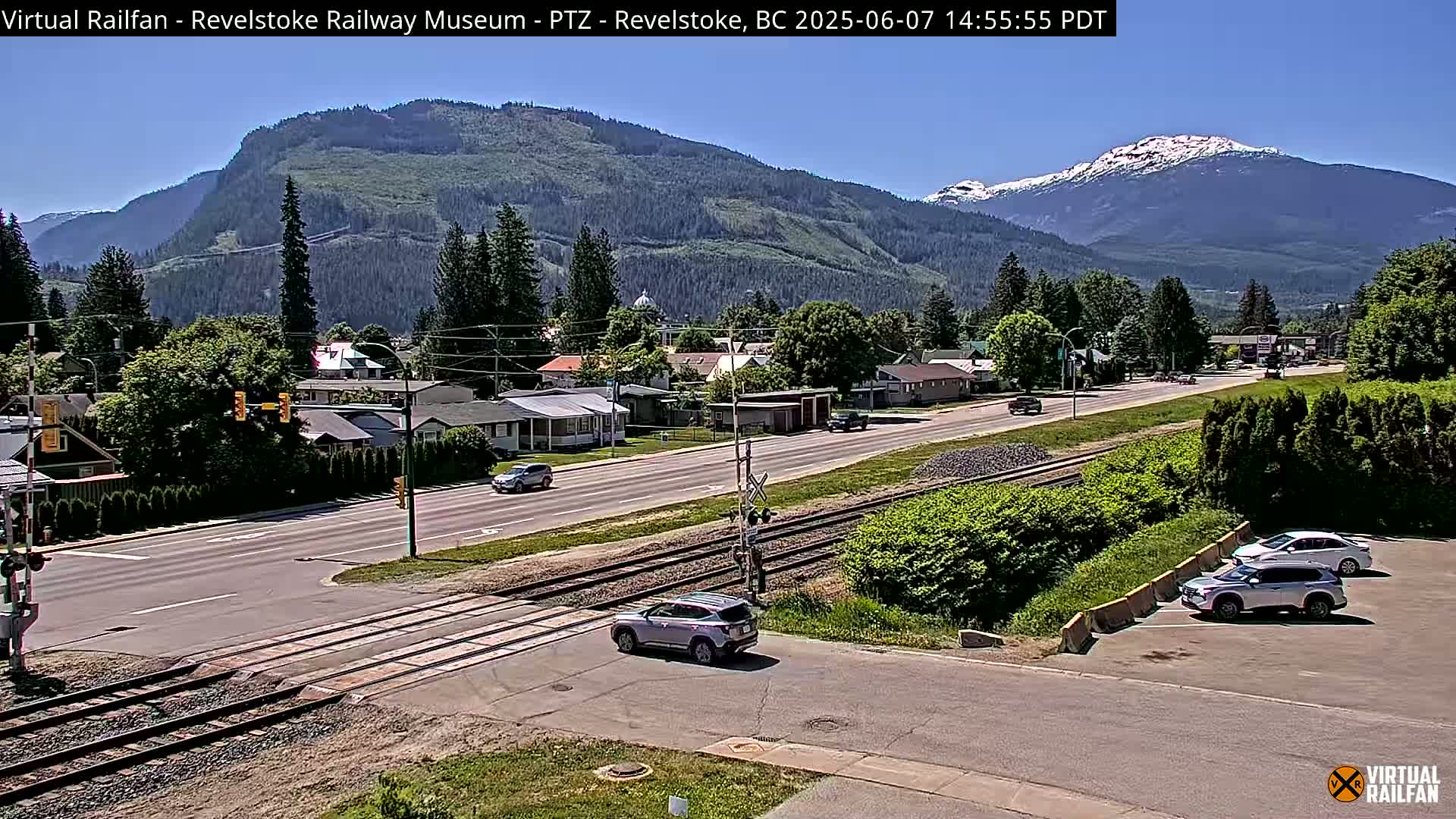 A sunny day shows a mountain range in the background of a small town with a roadway and train tracks crossing in the foreground, several cars are visible on the road and parked.