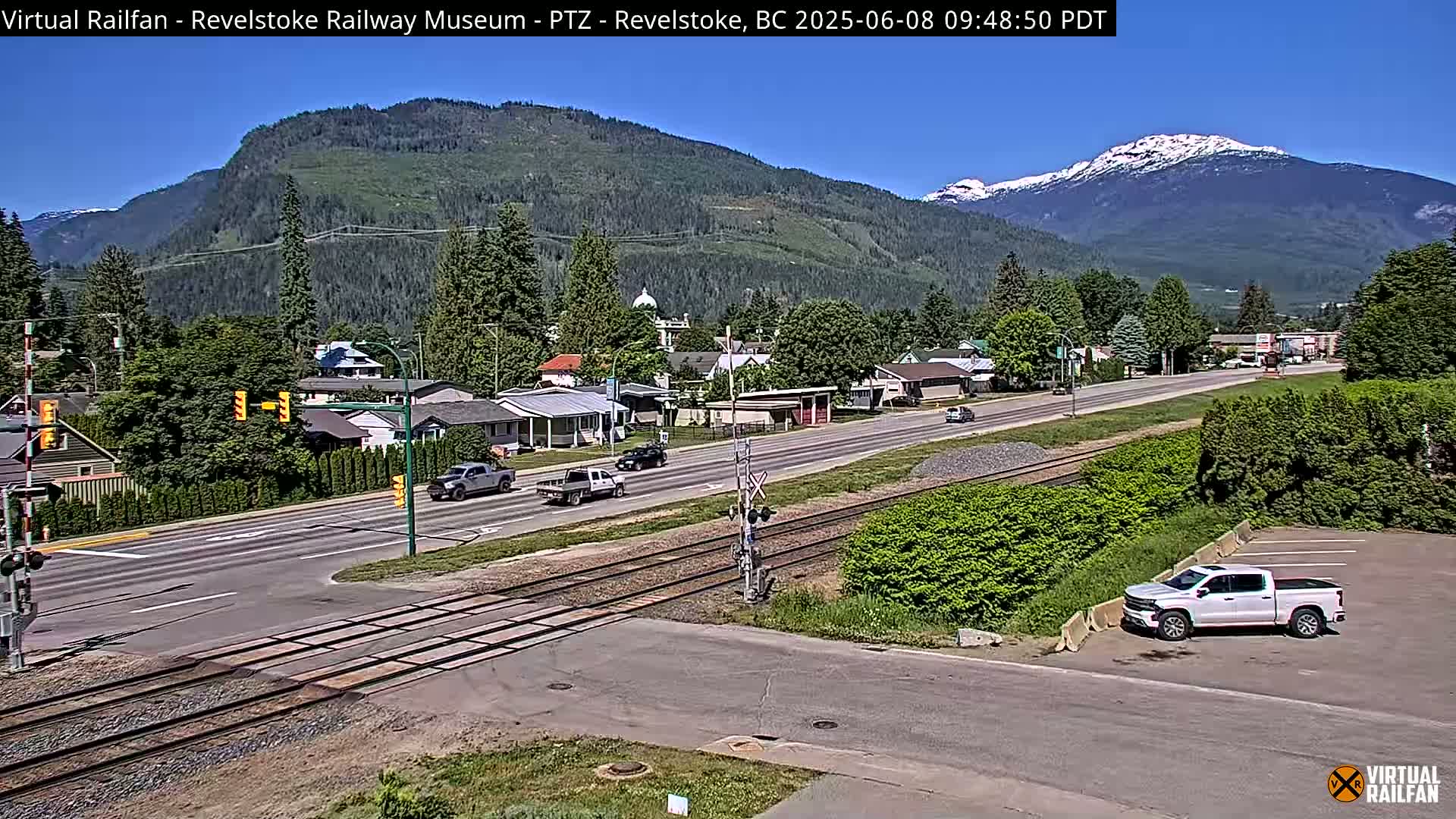 A sunny view of a town nestled in a valley between mountains, with a railway line crossing a road where several vehicles are driving.