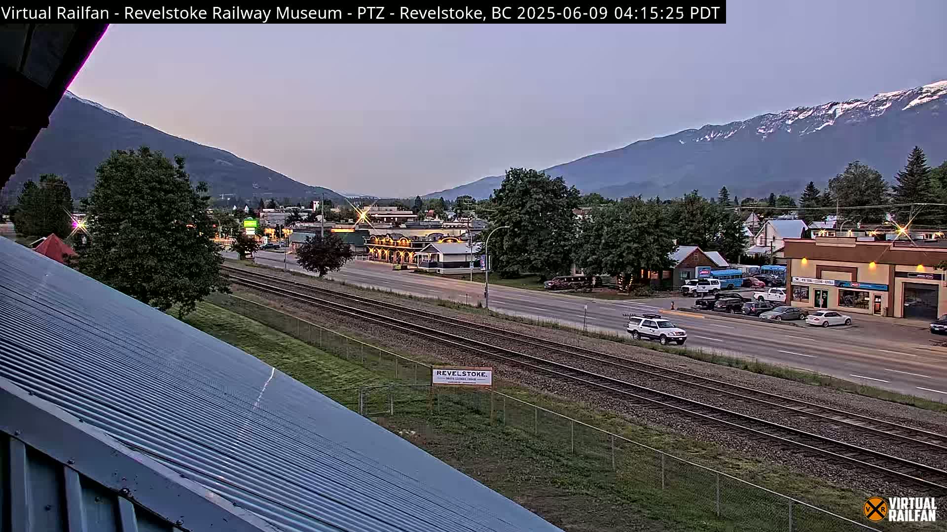 A long shot of a town at dusk, under a twilight sky, shows a railroad track next to a road, with mountains in the background.