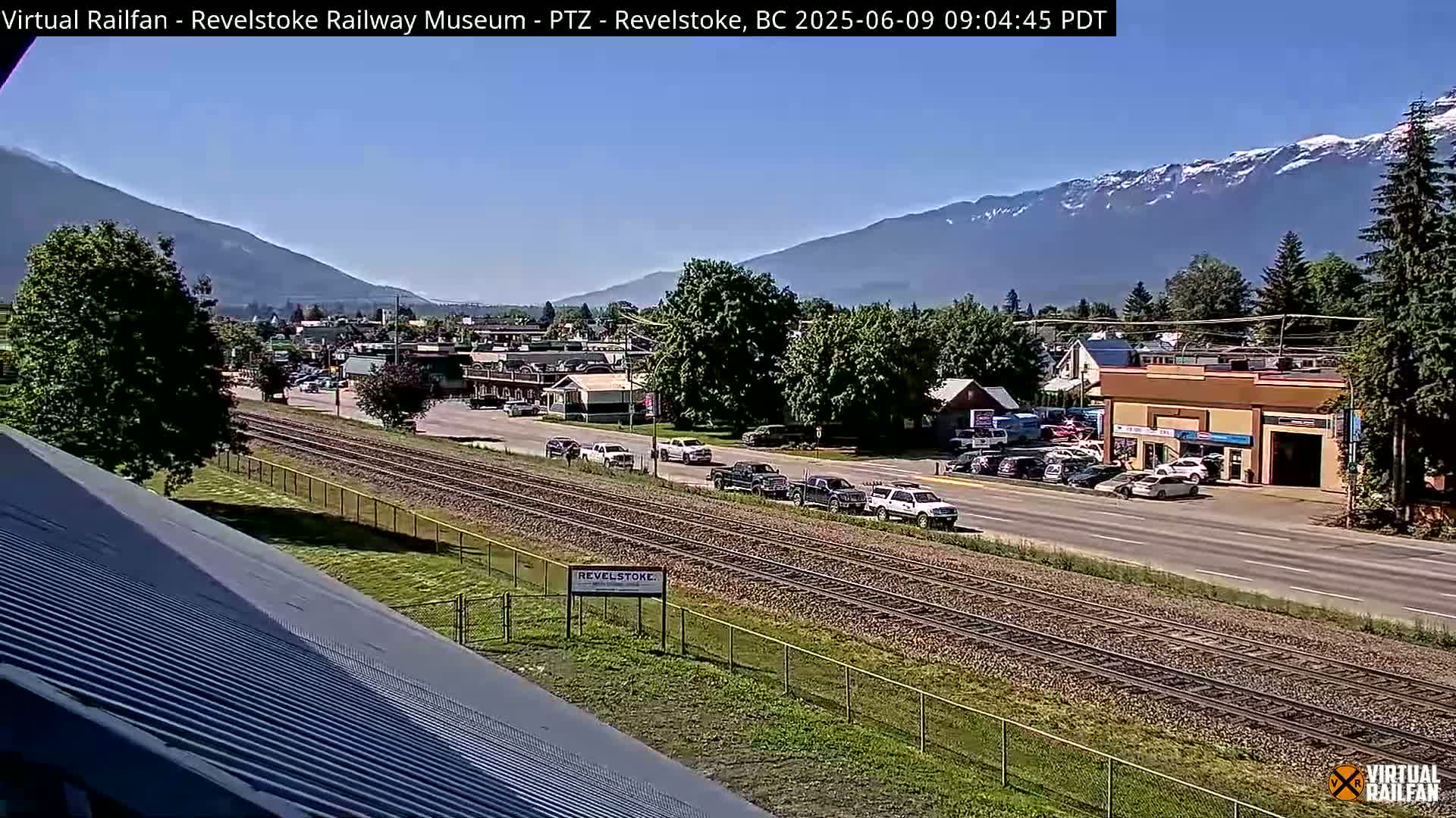 A sunny day reveals a town scene with a railroad track beside a road, several vehicles parked and driving, buildings, and mountains in the background.
