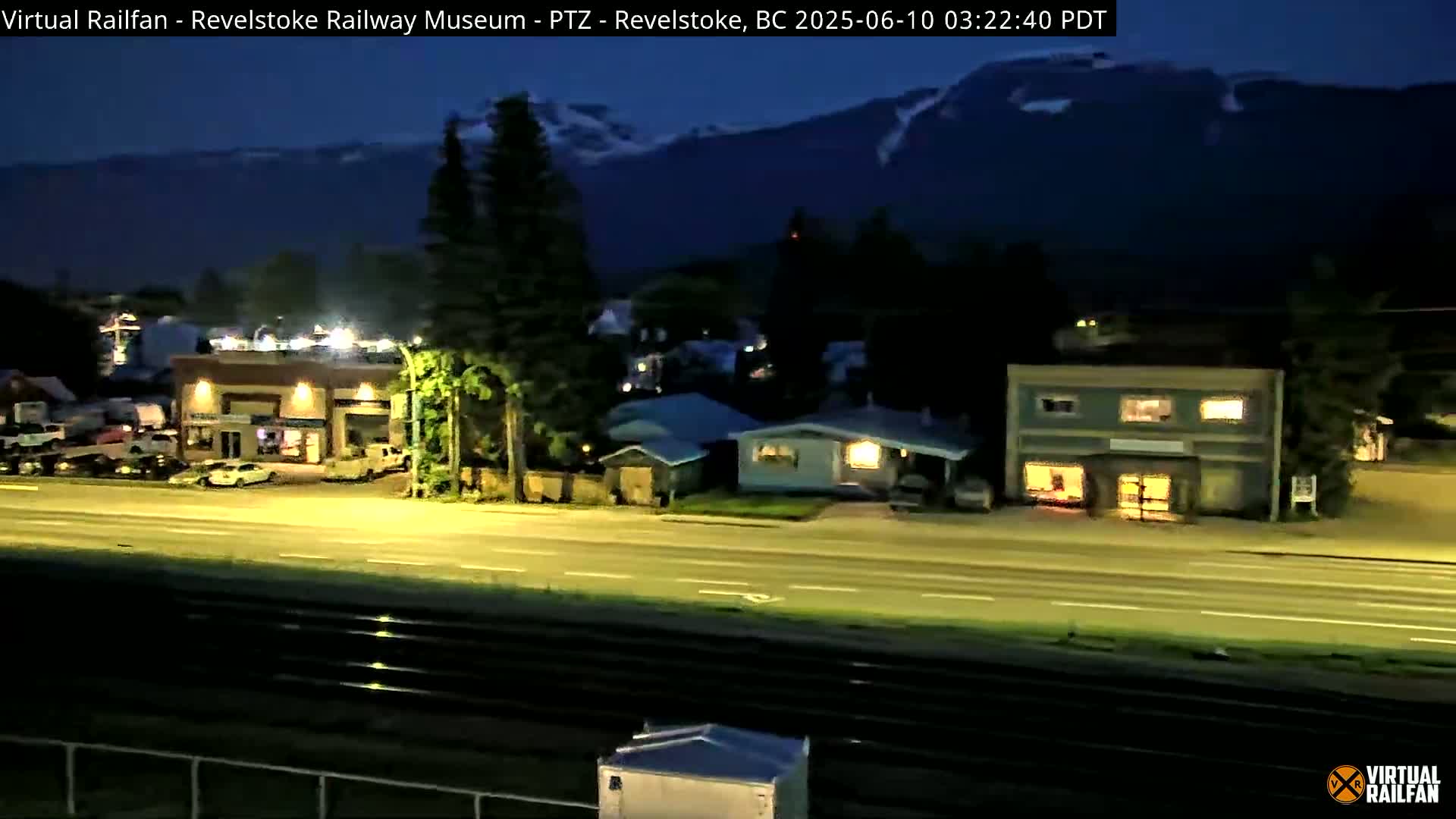 A nighttime view of a town with buildings and a road lit by streetlights, under a dark sky with mountains in the background.