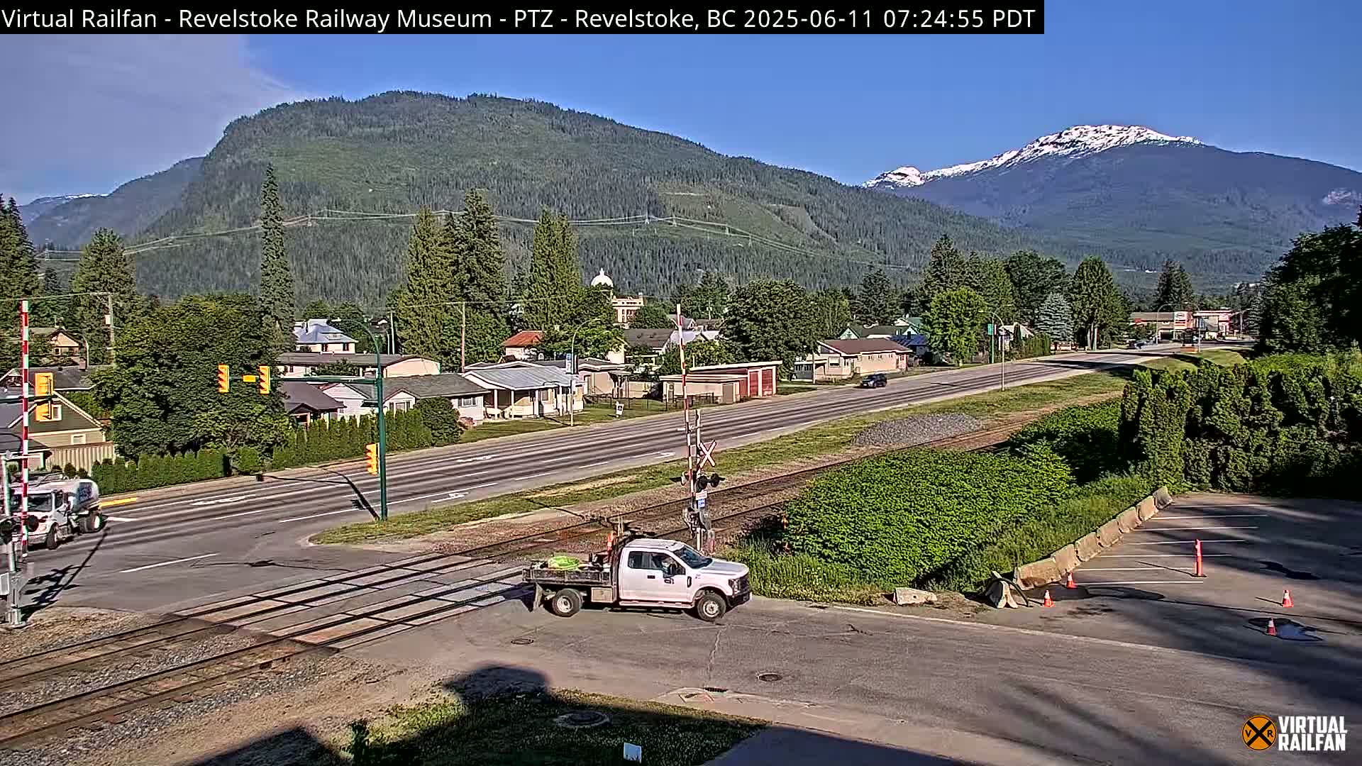 A sunny day reveals a view of a town nestled in a valley, with mountains in the background, some showing snow-capped peaks, and a roadway that crosses railroad tracks.
