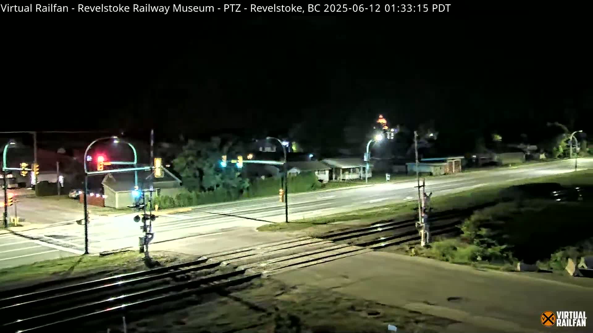 A nighttime view of a road intersecting railroad tracks at a traffic light, with sparse traffic and some streetlights illuminating the scene.