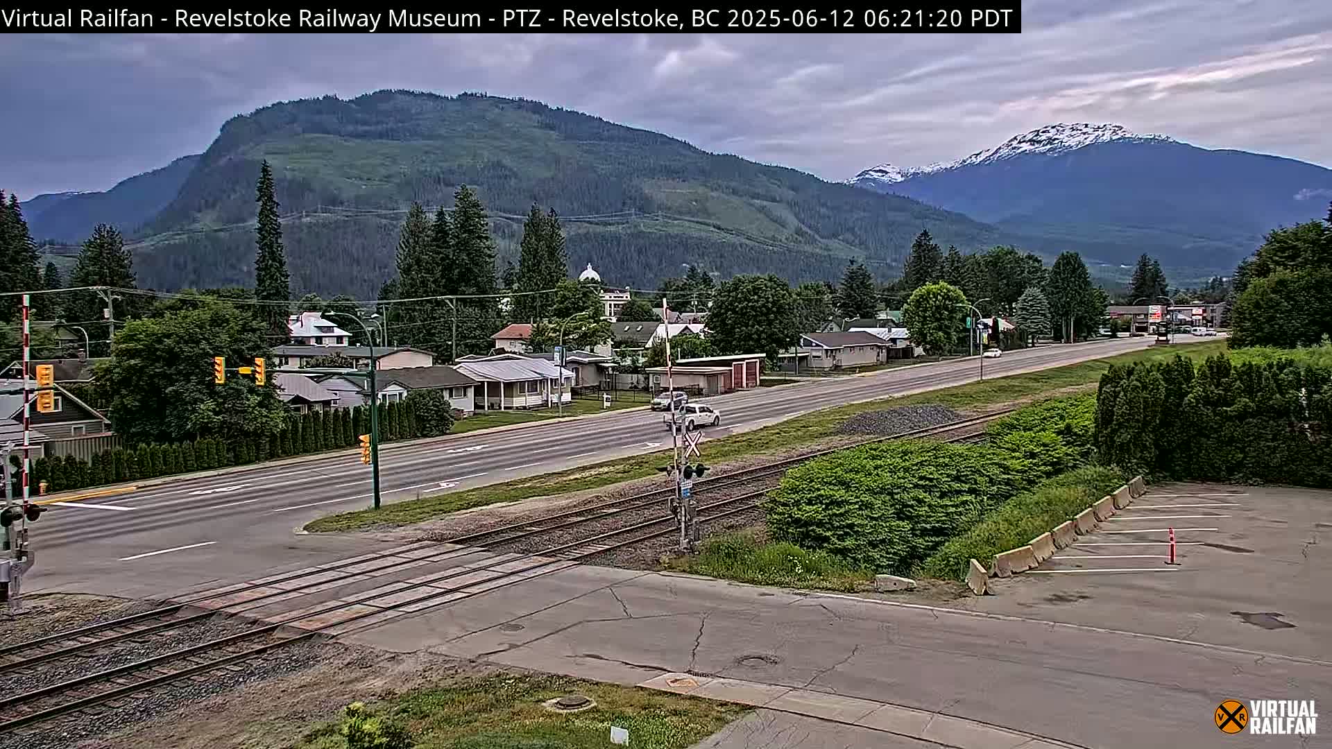 A partly cloudy view of a town with mountains in the background, showing a road crossing railroad tracks.
