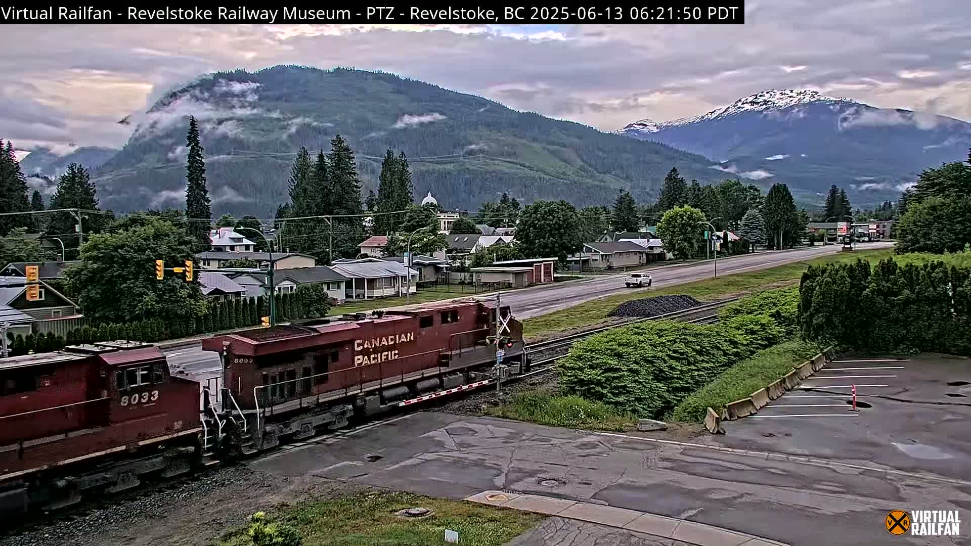A Canadian Pacific Railway train passes through a town nestled in a valley, with mountains shrouded in mist and clouds in the background.