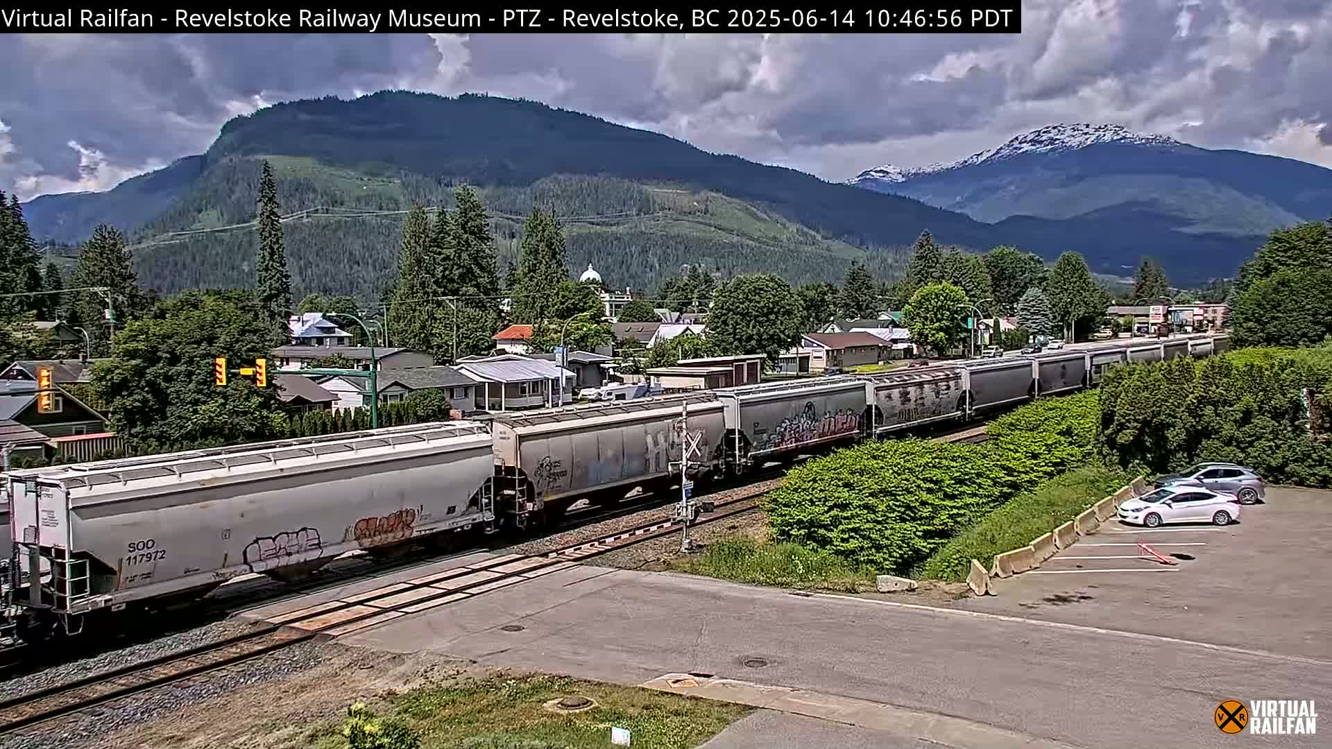 A long freight train passes through a residential area nestled in a valley beneath mountains on a partly cloudy day.