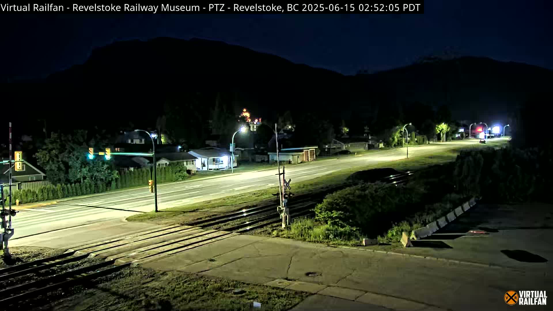 A nighttime view of a road and railway crossing in a small town, with dark mountains in the background under a clear, dark sky.