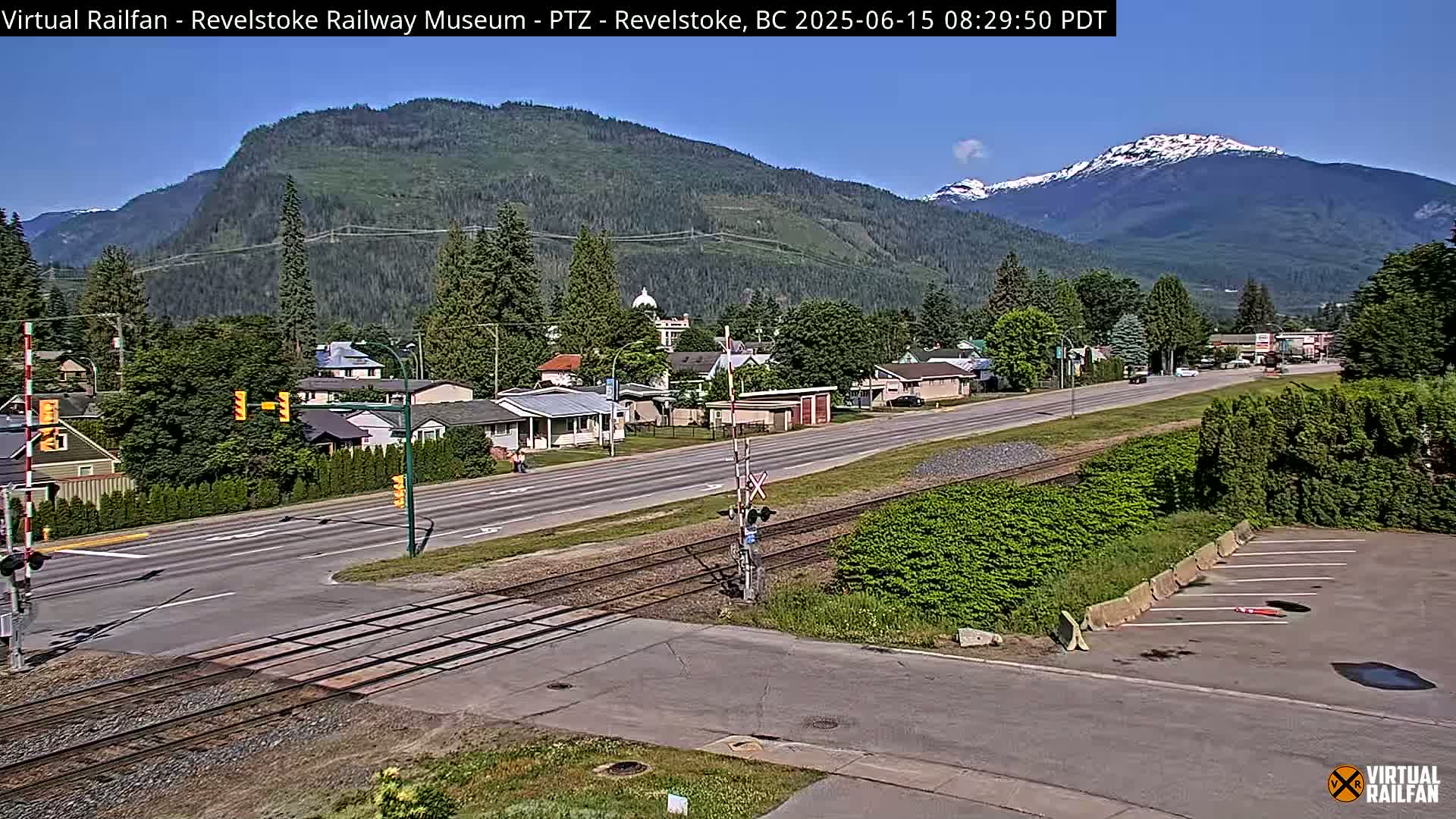 A sunny day reveals a town nestled at the base of a mountain range, with a railway crossing bisecting a road in the foreground.