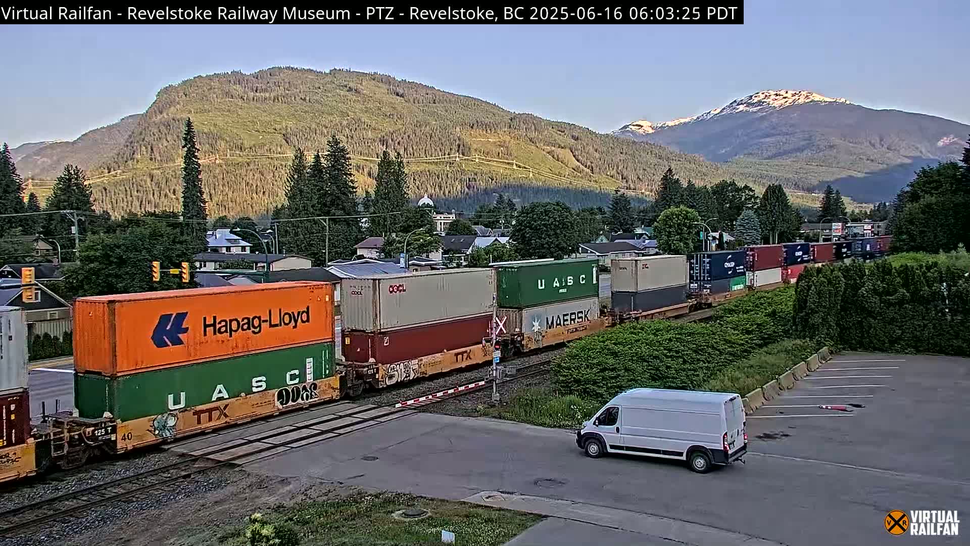 A long freight train carrying shipping containers passes through a town at the foot of a mountain range on a clear, sunny morning.