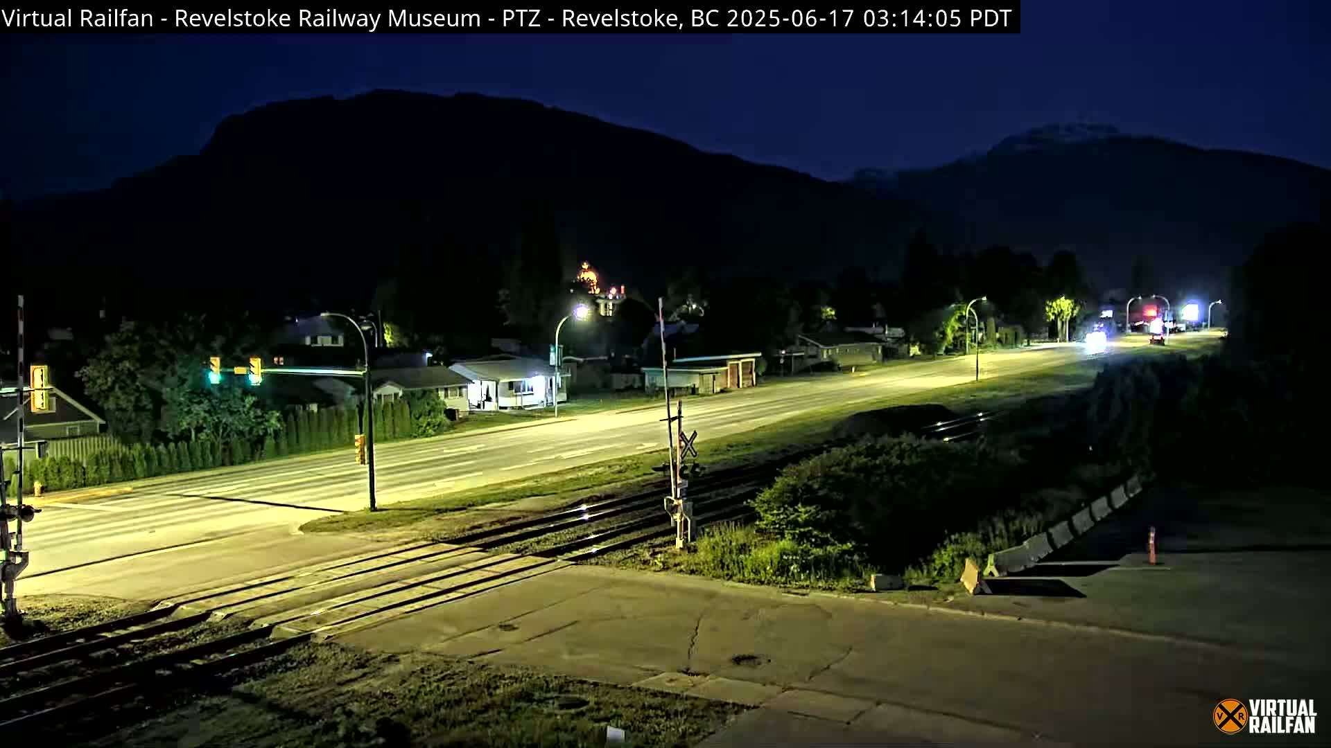 A nighttime view of a road and railway crossing, with a few vehicles on the road and a dark mountain range in the background under a clear, dark sky.