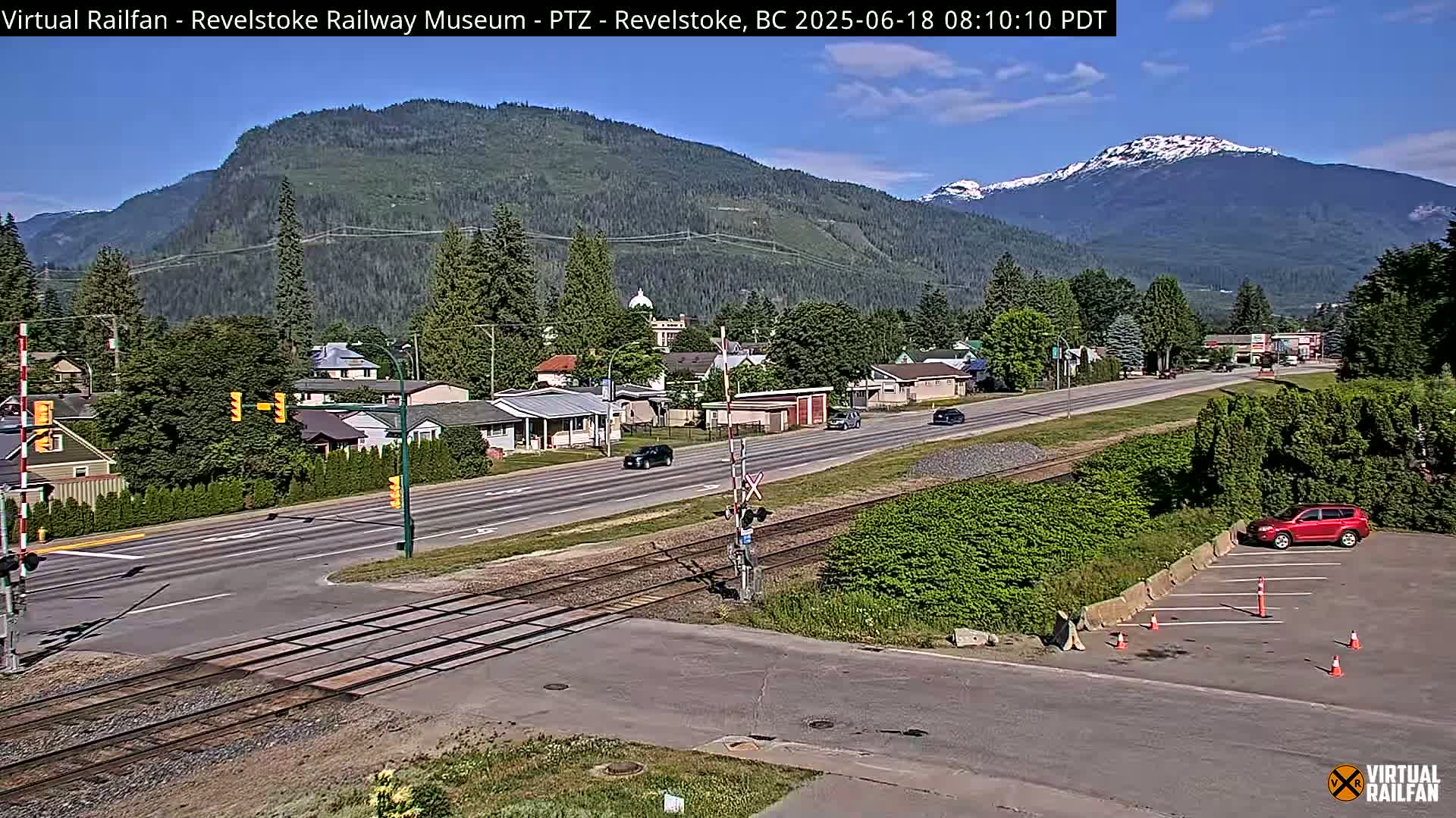 A sunny day reveals a town nestled in a valley, with mountains in the background, some showing snow, alongside railroad tracks that cross a road.