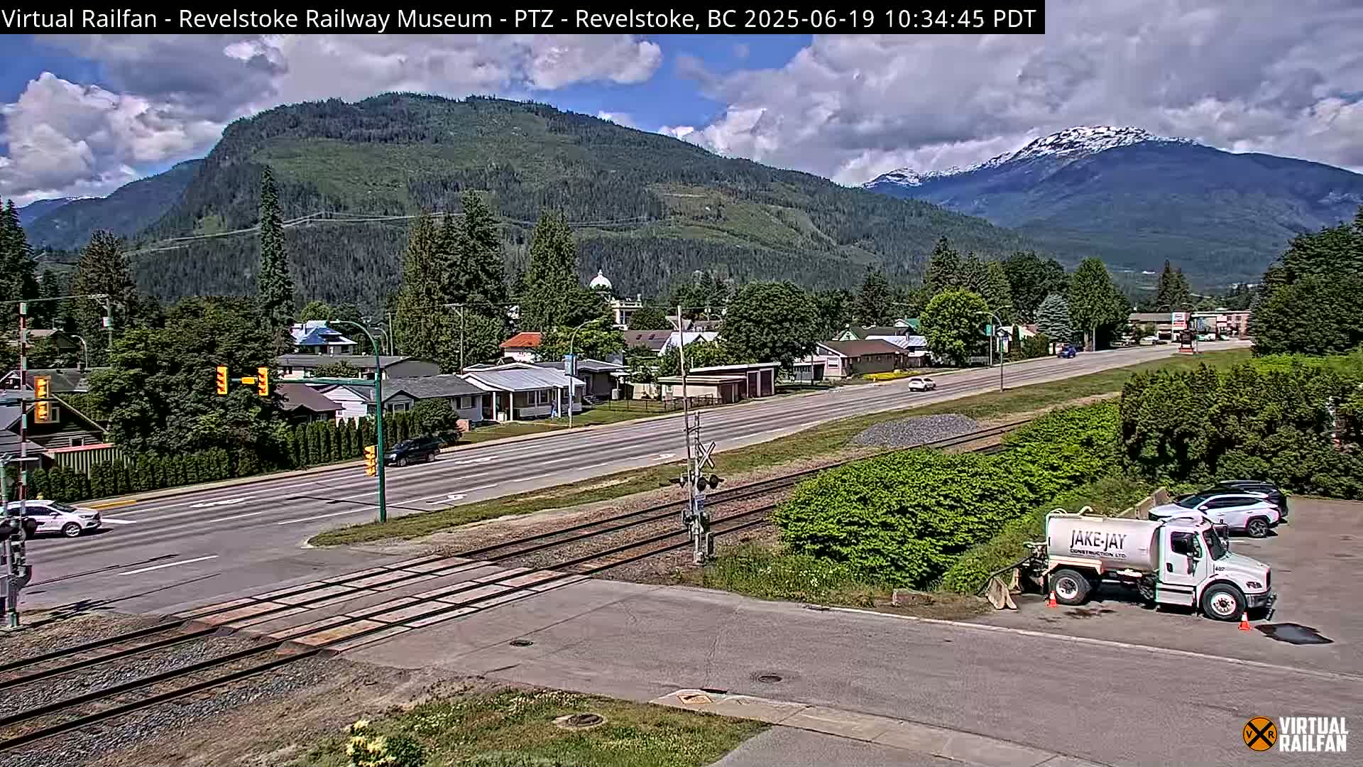 A partly cloudy day reveals a town nestled in a valley between mountains, one of which has a snow-capped peak; railroad tracks cross a road in the foreground.