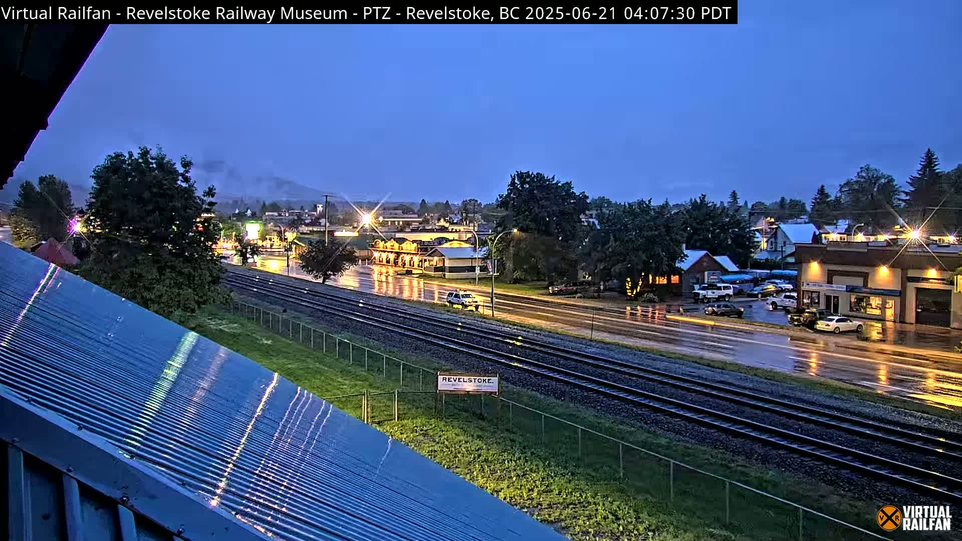 A wet, nighttime view of a town with train tracks running through it, seen from a building's corrugated metal roof.