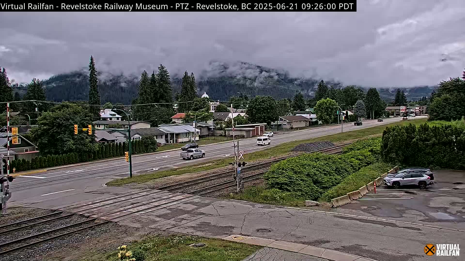 A cloudy day reveals a town with a railroad crossing, several vehicles on the road and in a parking lot, and a mountain range in the background partially obscured by clouds.
