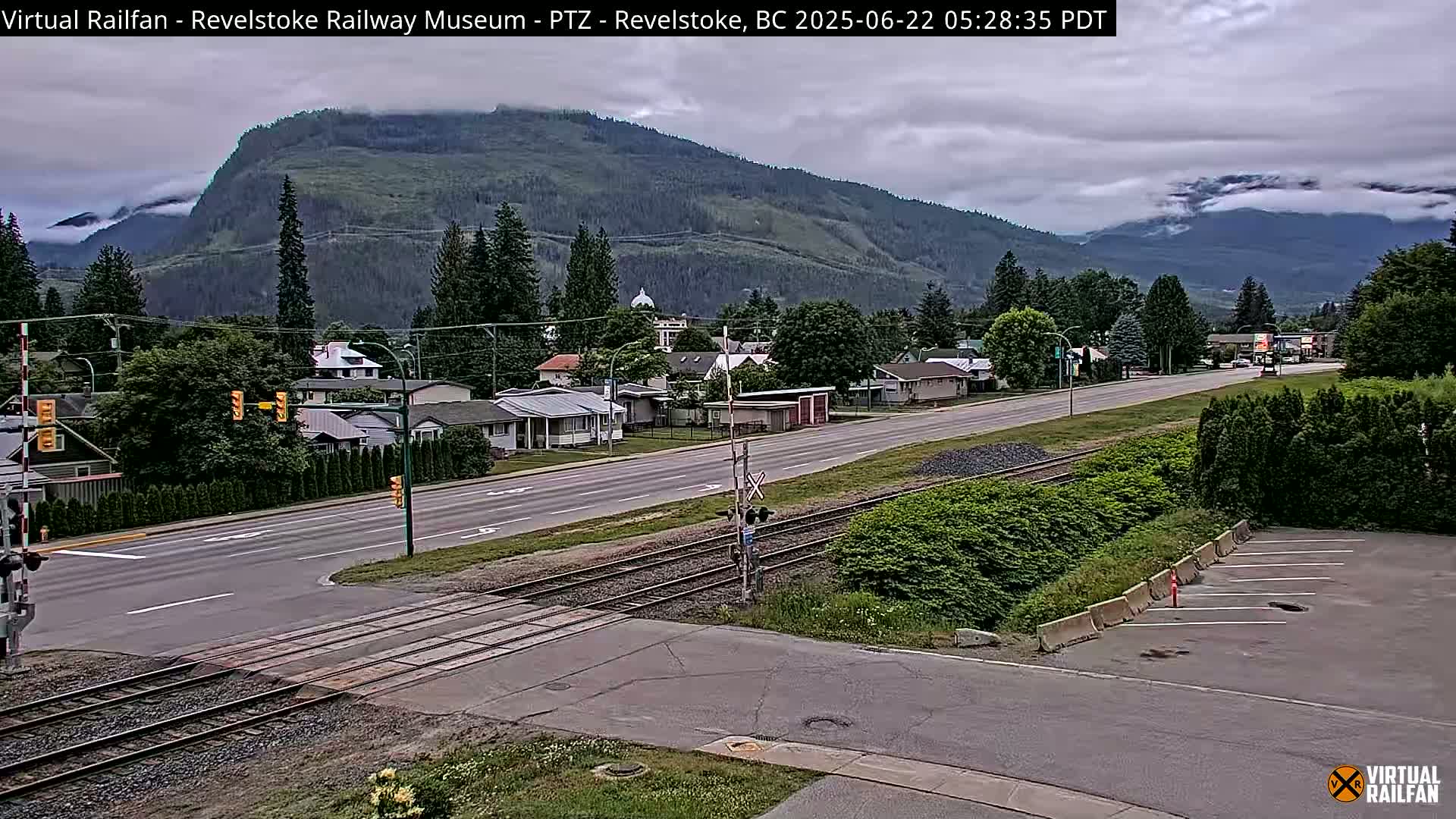 A mostly cloudy day reveals a town nestled at the base of a mountain range, with train tracks crossing a road in the foreground.