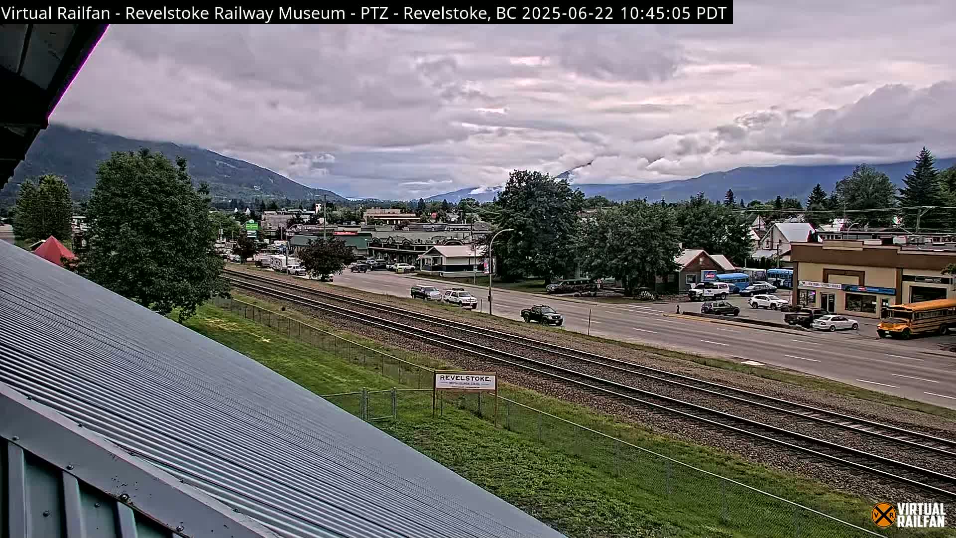 A partly cloudy day reveals a view of a town nestled in a valley between mountains, with railroad tracks running through the foreground.