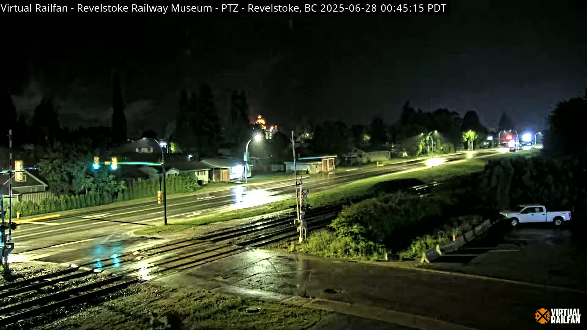 A rain-slicked nighttime street scene shows a railroad crossing with vehicles and streetlights illuminating wet roadways and surrounding vegetation.