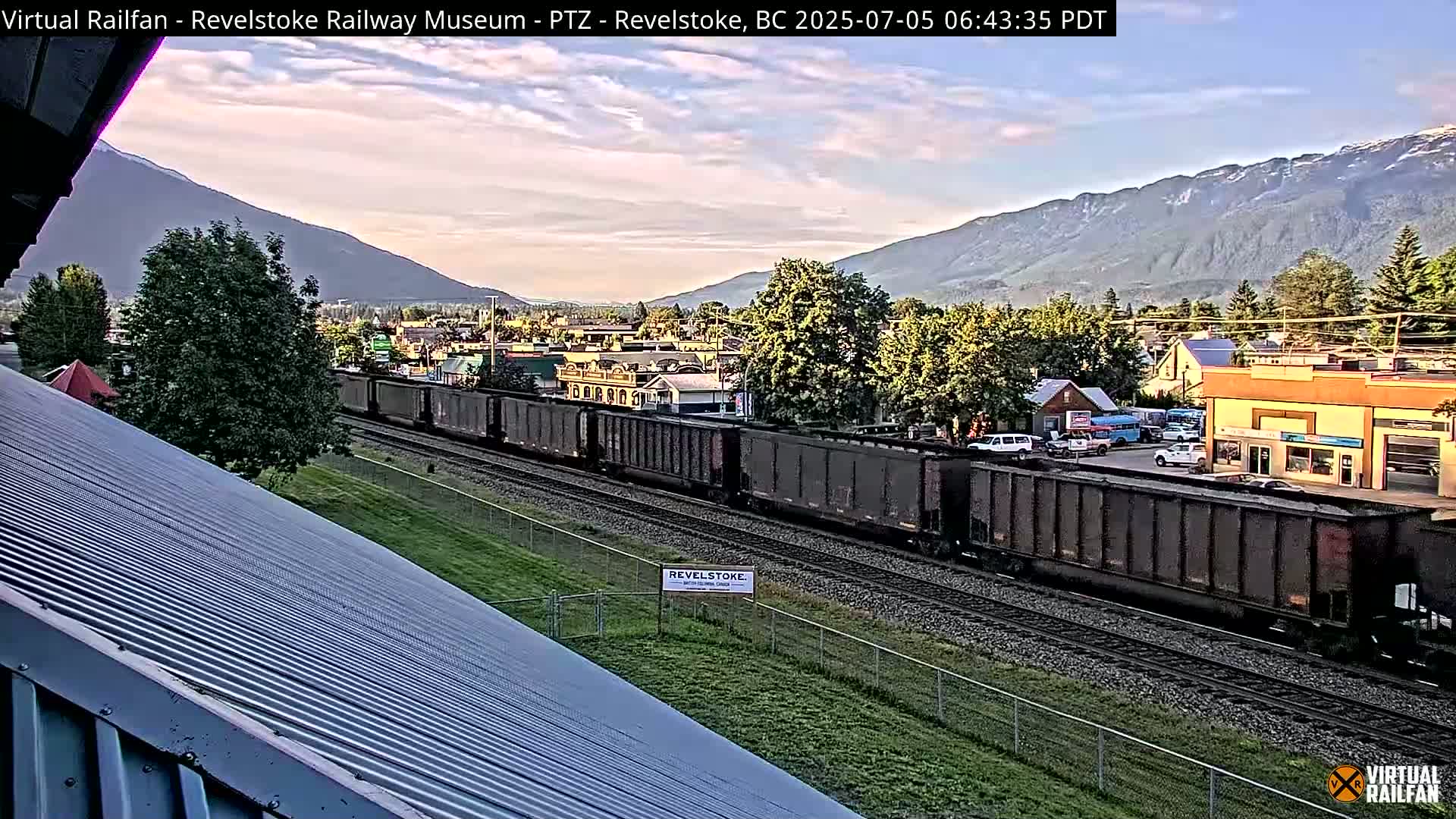 A long freight train travels along railroad tracks past a small town nestled in a valley beneath mountains, under a partly cloudy sky.