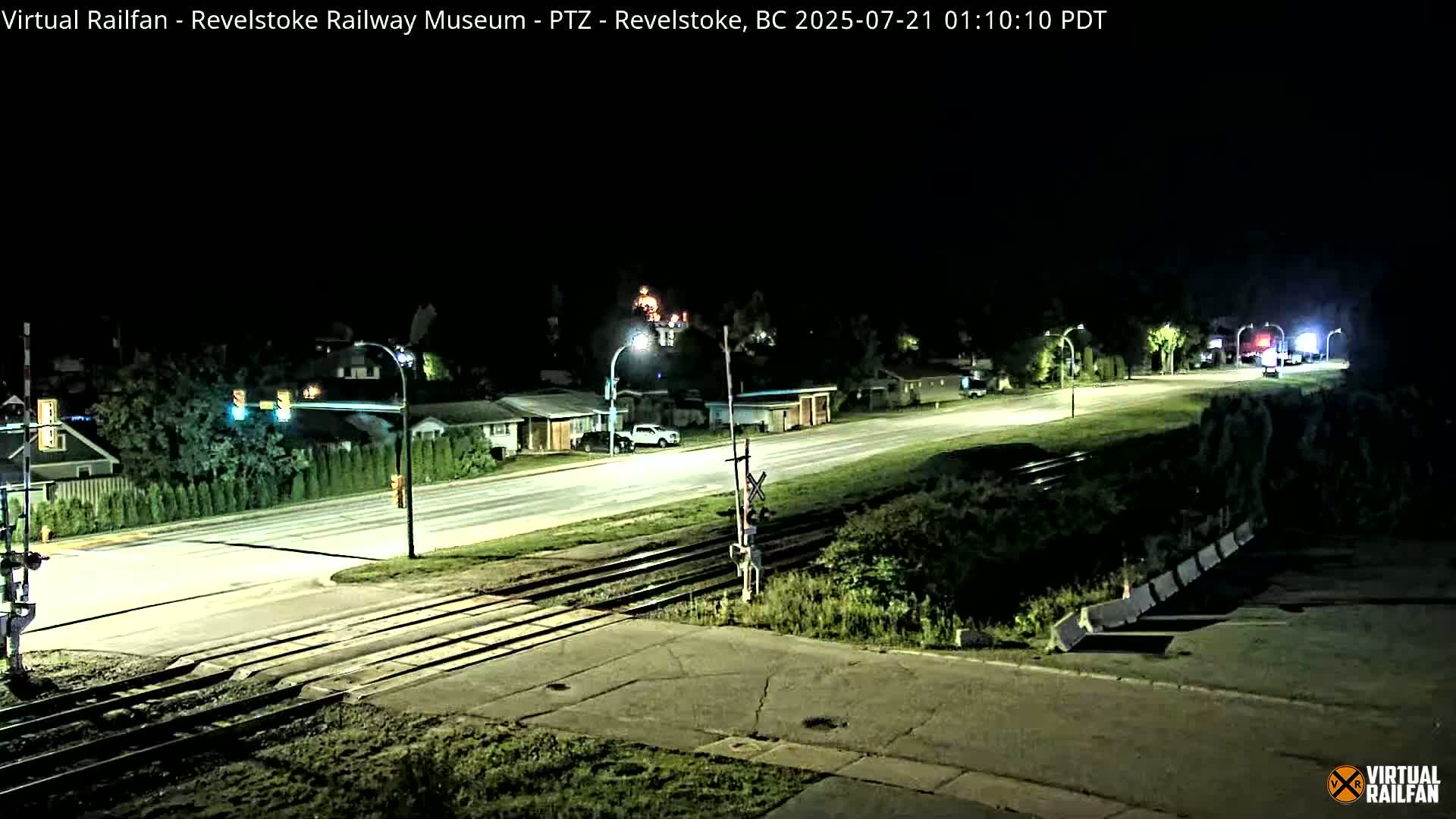 A nighttime view of a mostly empty road intersecting with train tracks, illuminated by streetlights, with a few vehicles visible in the distance.