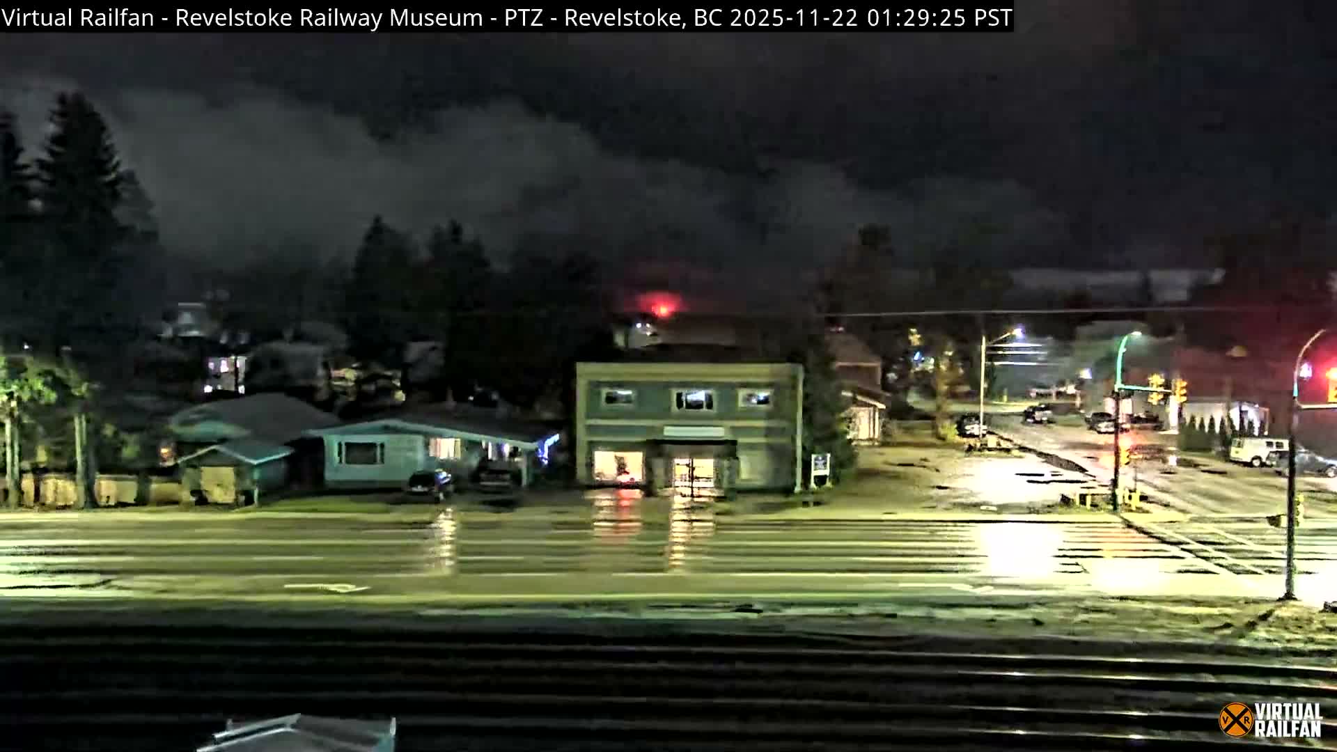 A wet town street with commercial and residential buildings, illuminated by artificial lights and featuring train tracks in the foreground, is seen at night under rainy conditions.