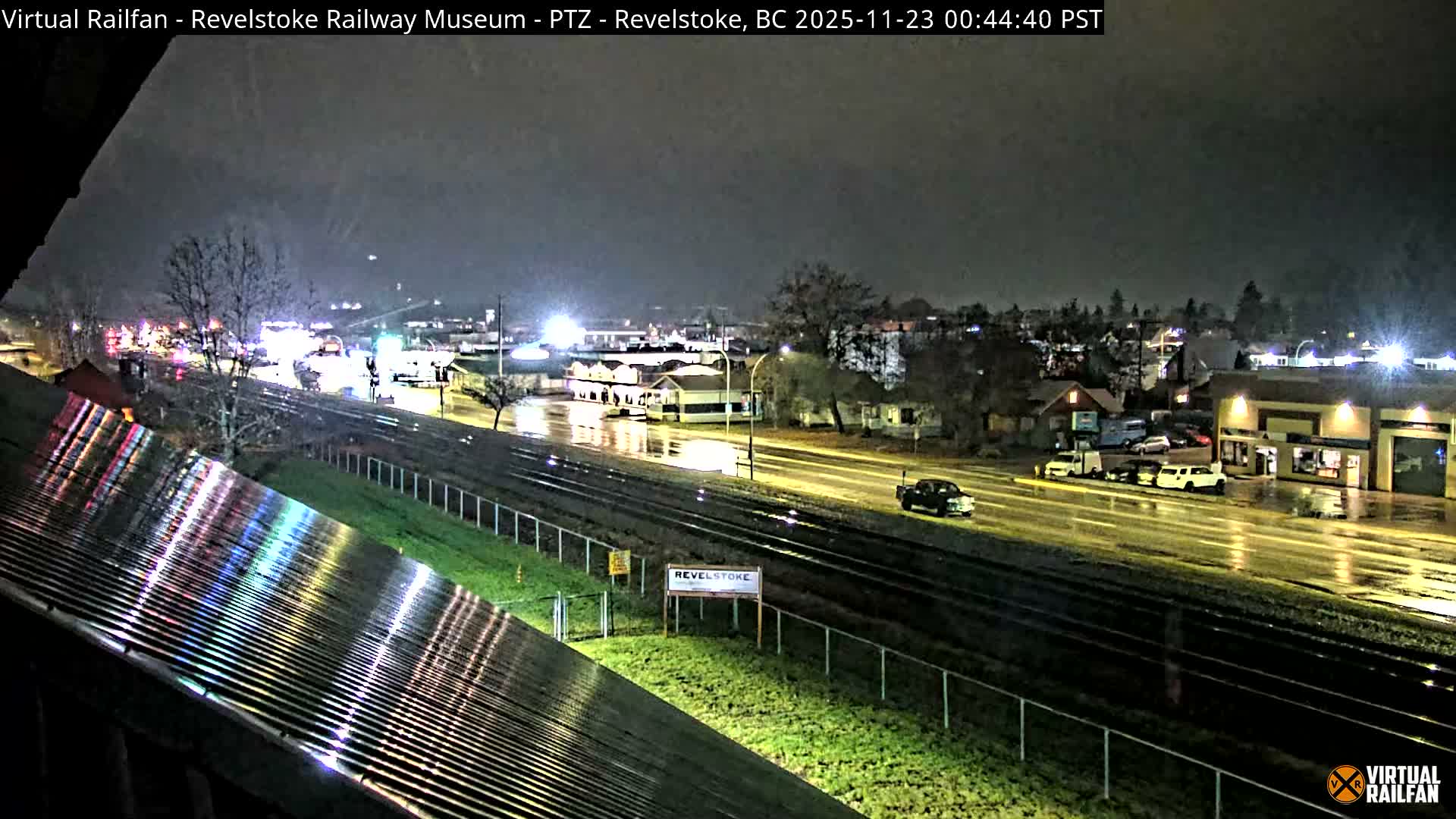 The image captures a rainy night scene in an urban area, with wet roads and buildings illuminated by streetlights, alongside railway tracks, under a dark, overcast sky.