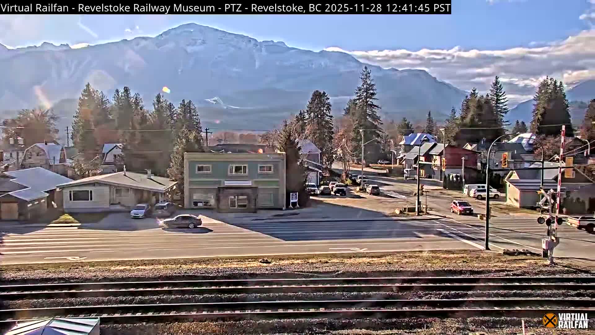 A sunny day with some white clouds illuminates a town nestled among evergreen trees and snow-capped mountains, with railroad tracks in the foreground, a street with vehicles and buildings in the midground, and a railway crossing barrier on the right.