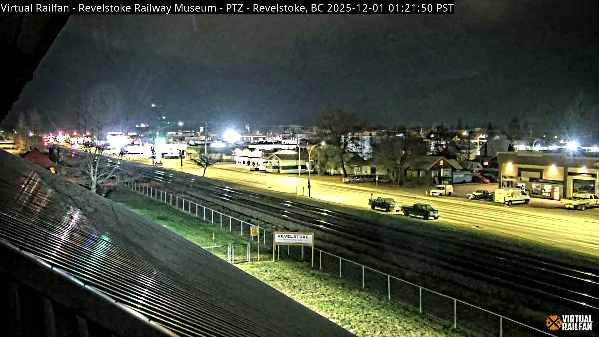A clear, dark night reveals a town scene featuring brightly lit streets, buildings, and vehicles running alongside multiple railroad tracks, with wet surfaces reflecting the artificial lights.