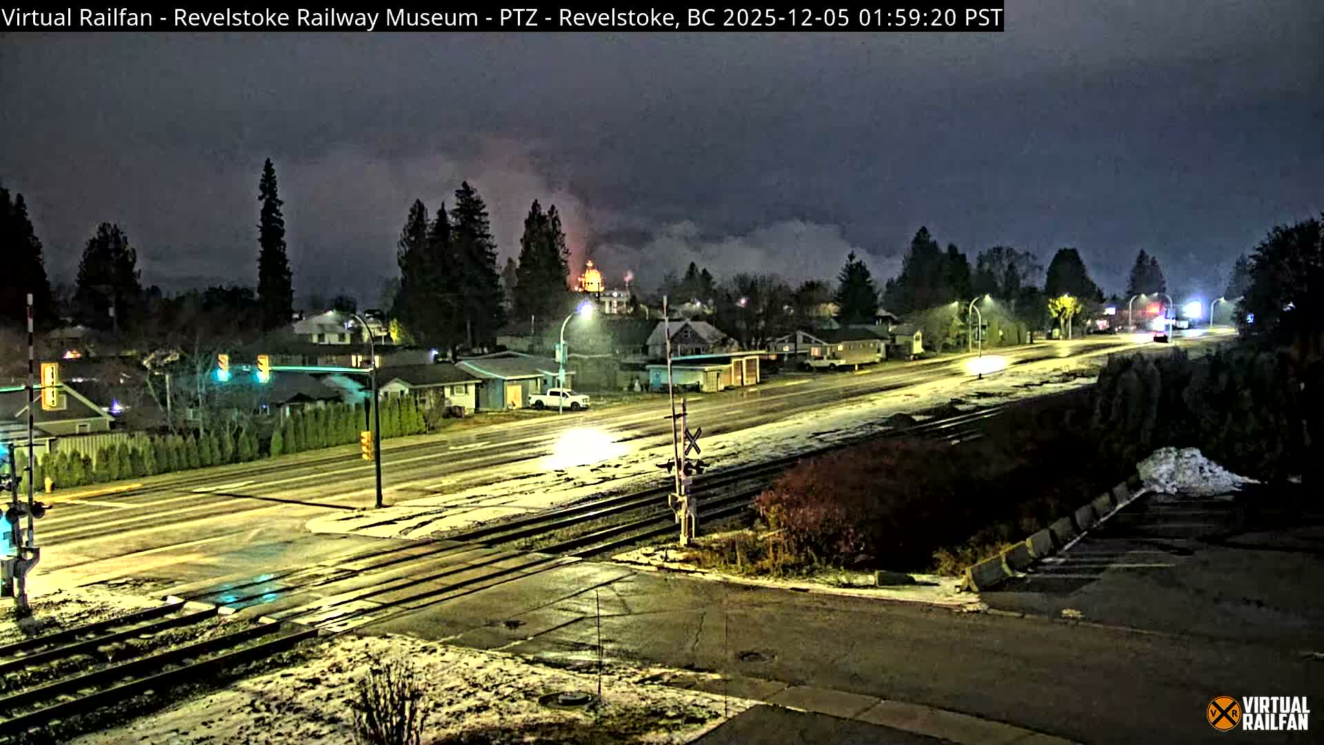 The image captures a nighttime view of a town street with a light dusting of snow covering the ground, featuring railroad tracks crossing an intersection, houses illuminated by streetlights, and a distant building with a glowing dome emitting steam under a cloudy sky.