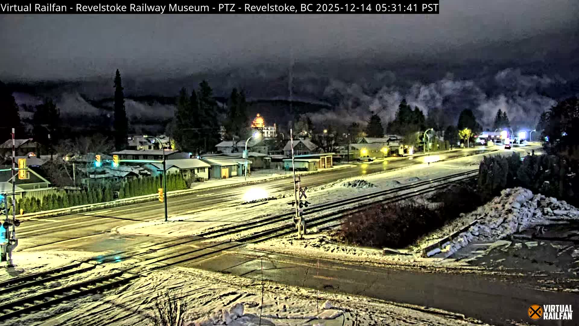 The image captures a nighttime view of a town street with a light dusting of snow covering the ground, featuring railroad tracks crossing an intersection, houses illuminated by streetlights, and a distant building with a glowing dome emitting steam under a cloudy sky.
