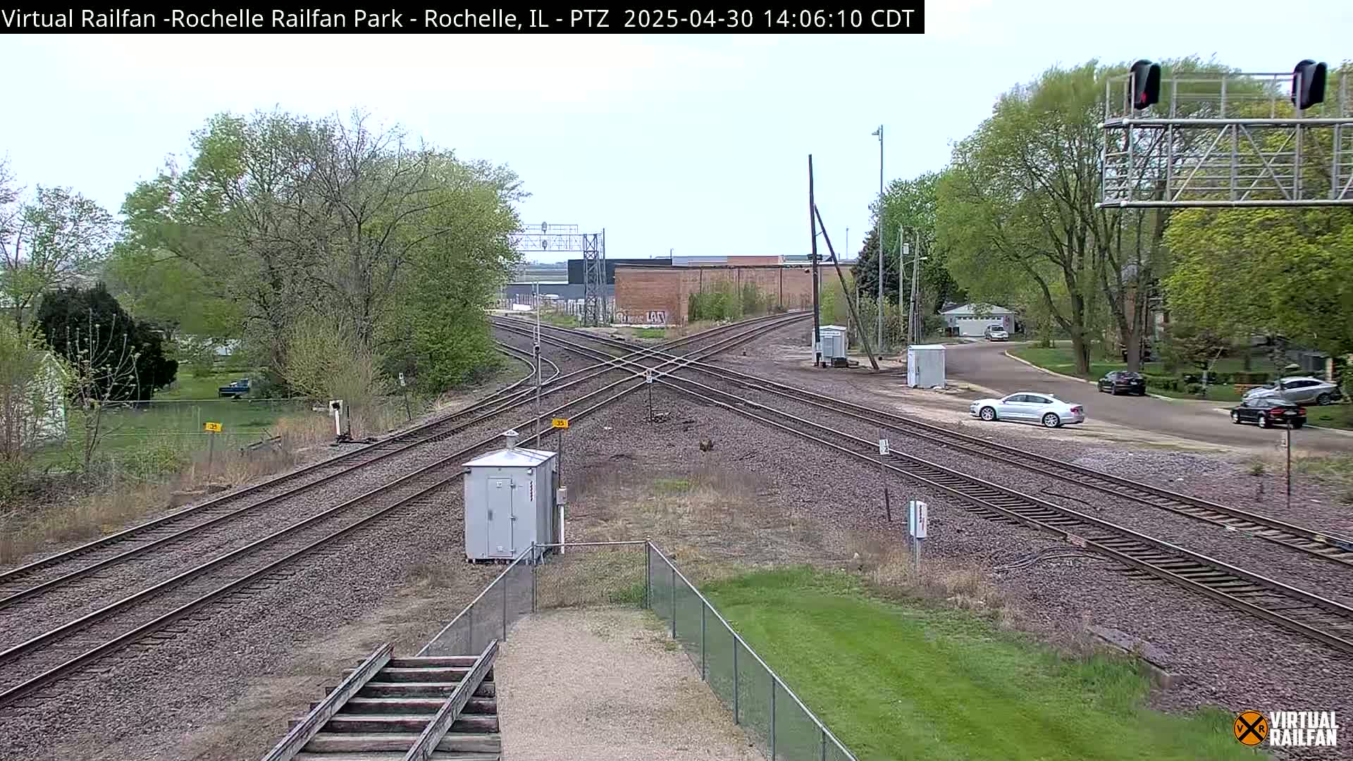 A sunny day shows a train crossing with several tracks and a few cars parked nearby, surrounded by green grass and trees.